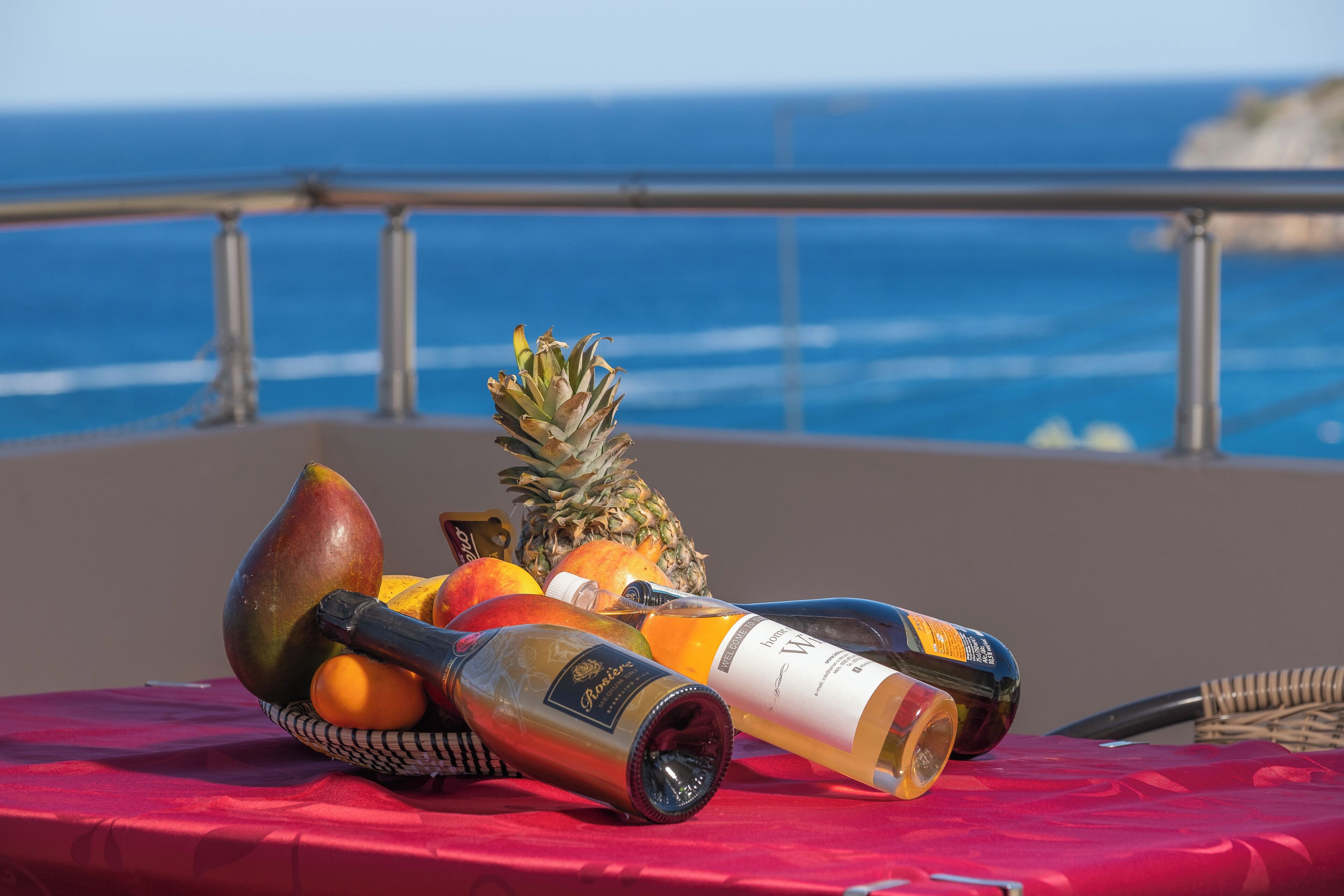 Una mesa con frutas y botellas sobre un mantel rojo, con vista al océano.