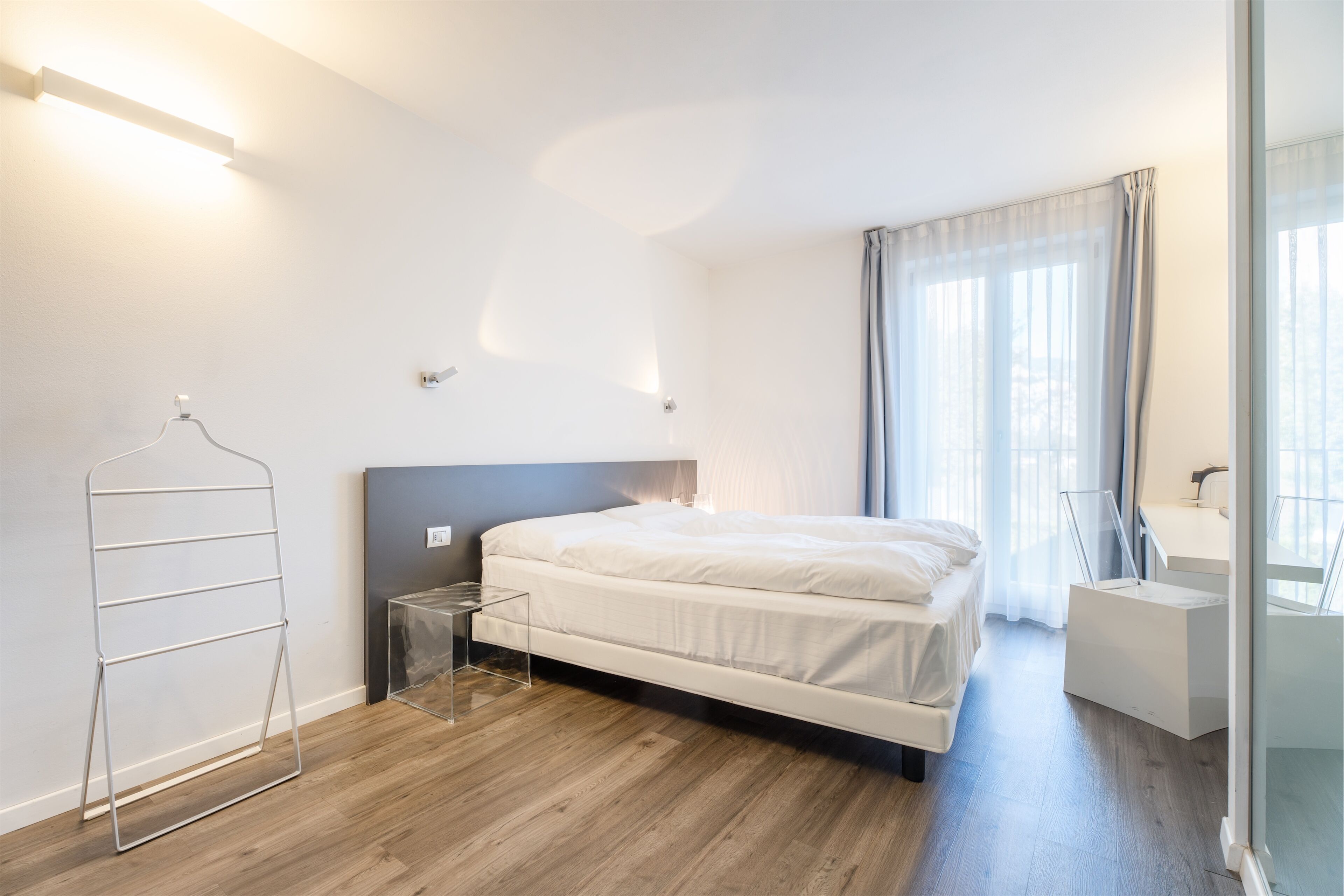 A modern bedroom with a bed, a bedside table, a hanging rack, and a bathroom with a mirror and sink.