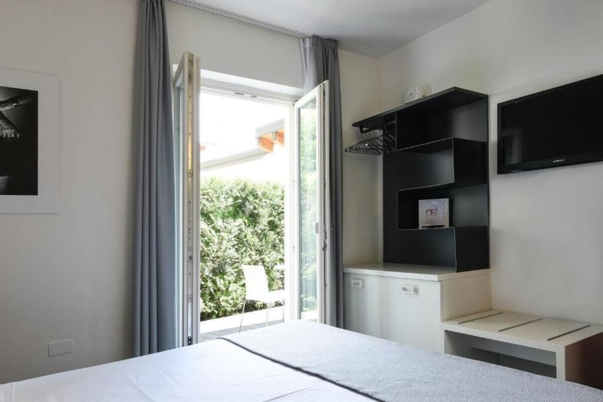 Ein Hotelzimmer mit einem Bett, einem an der Wand montierten Fernseher, einem kleinen Schreibtisch und einem Fenster mit Vorhängen.