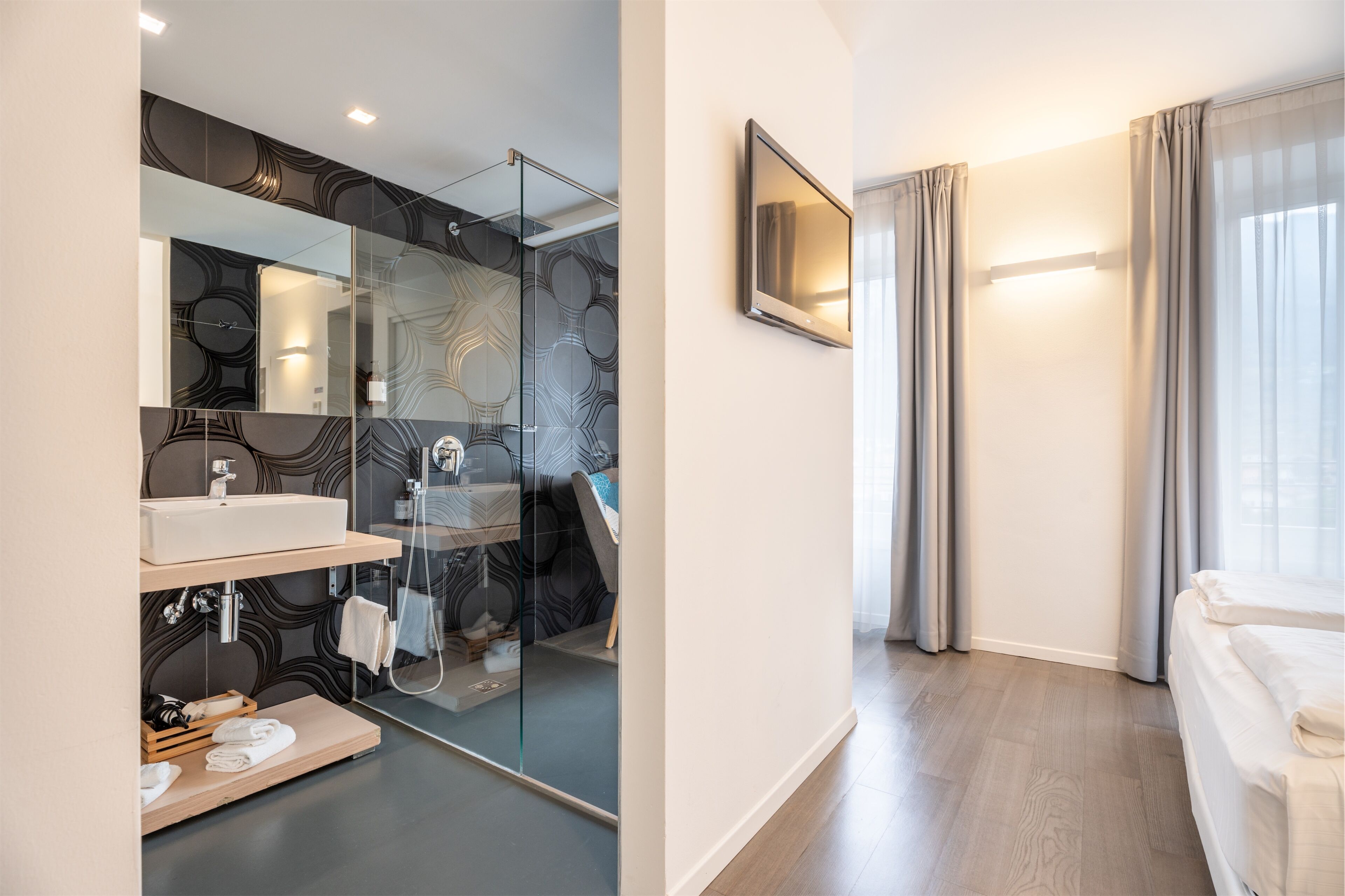 Un baño moderno con cabina de ducha de vidrio, un lavamanos blanco y un espejo. El cuarto tiene piso de madera y un ventanal grande con cortinas.