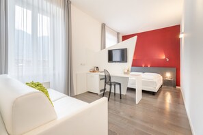 A modern hotel room with a white sofa, a desk with a chair, a bed, a TV mounted on the wall, and a red accent wall. - Garnì On The Rock (Arco)