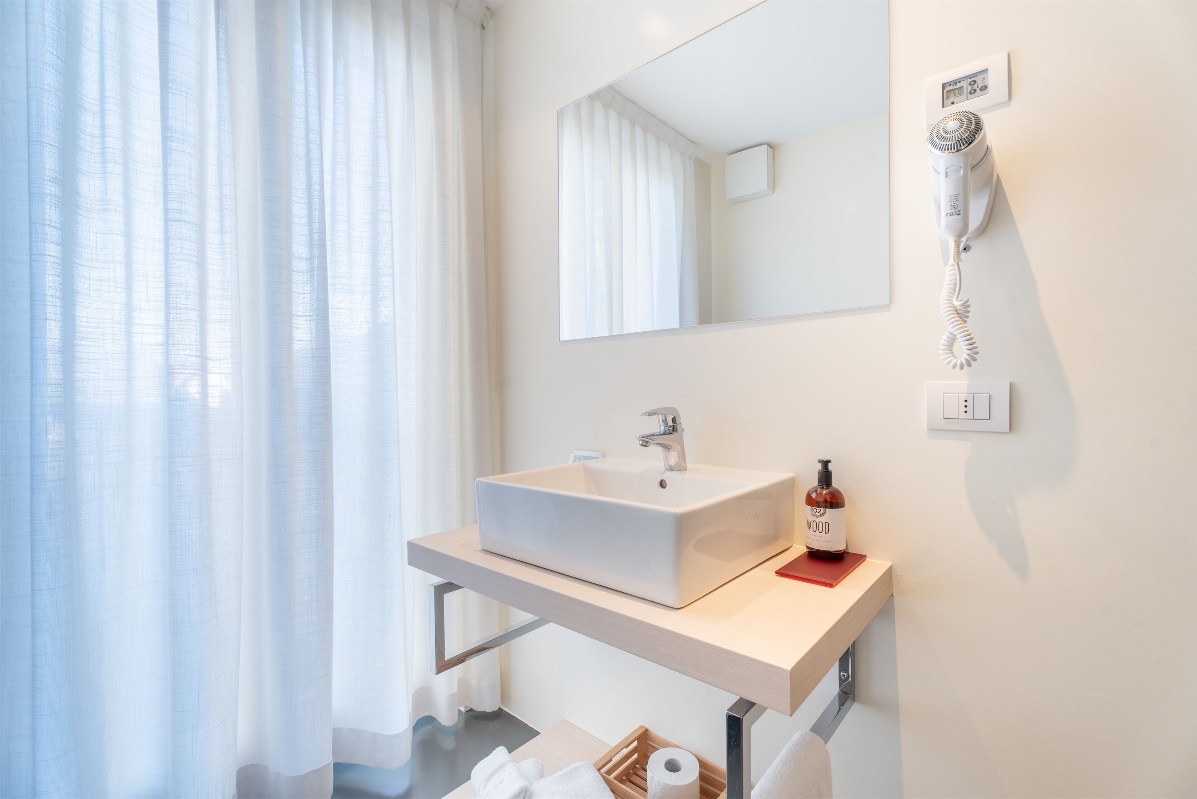 A modern bathroom with a white sink, a mirror, a hairdryer mounted on the wall, and a bottle of hand soap.