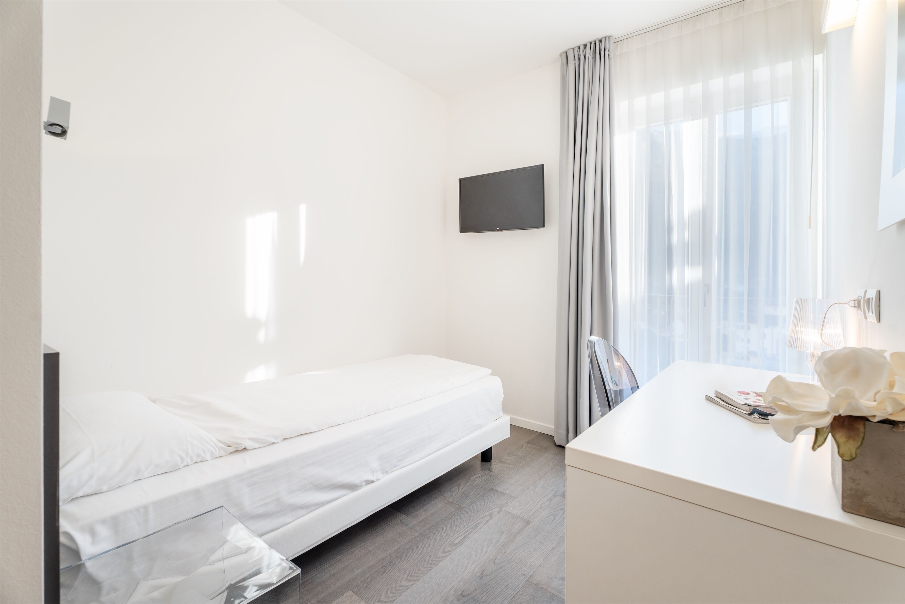 A modern hotel room with a single bed, a flat-screen TV mounted on the wall, a small desk with a vase of flowers, and a window with sheer curtains.