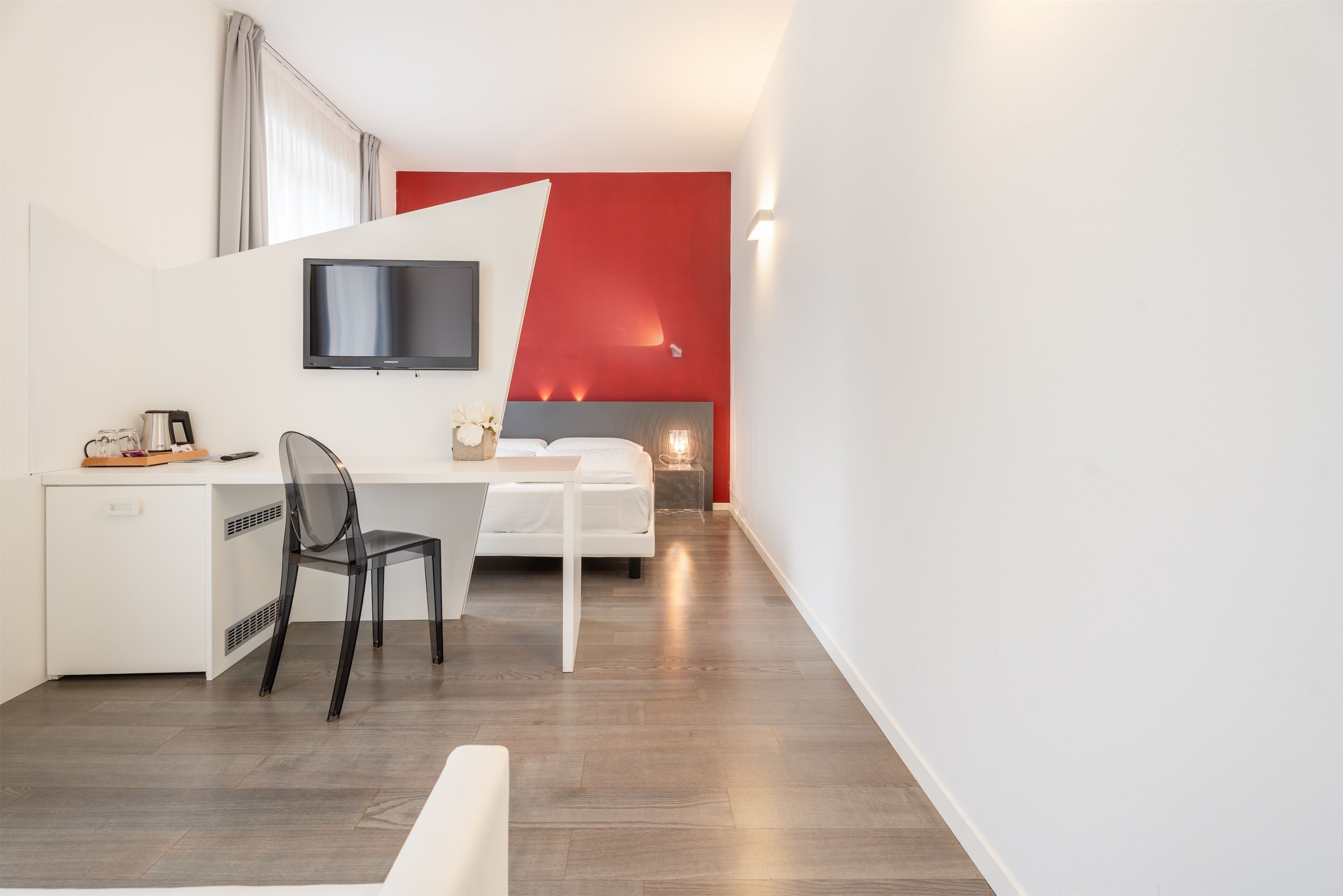 A modern hotel room with a bed, a desk, a chair, a TV, and a red accent wall.
