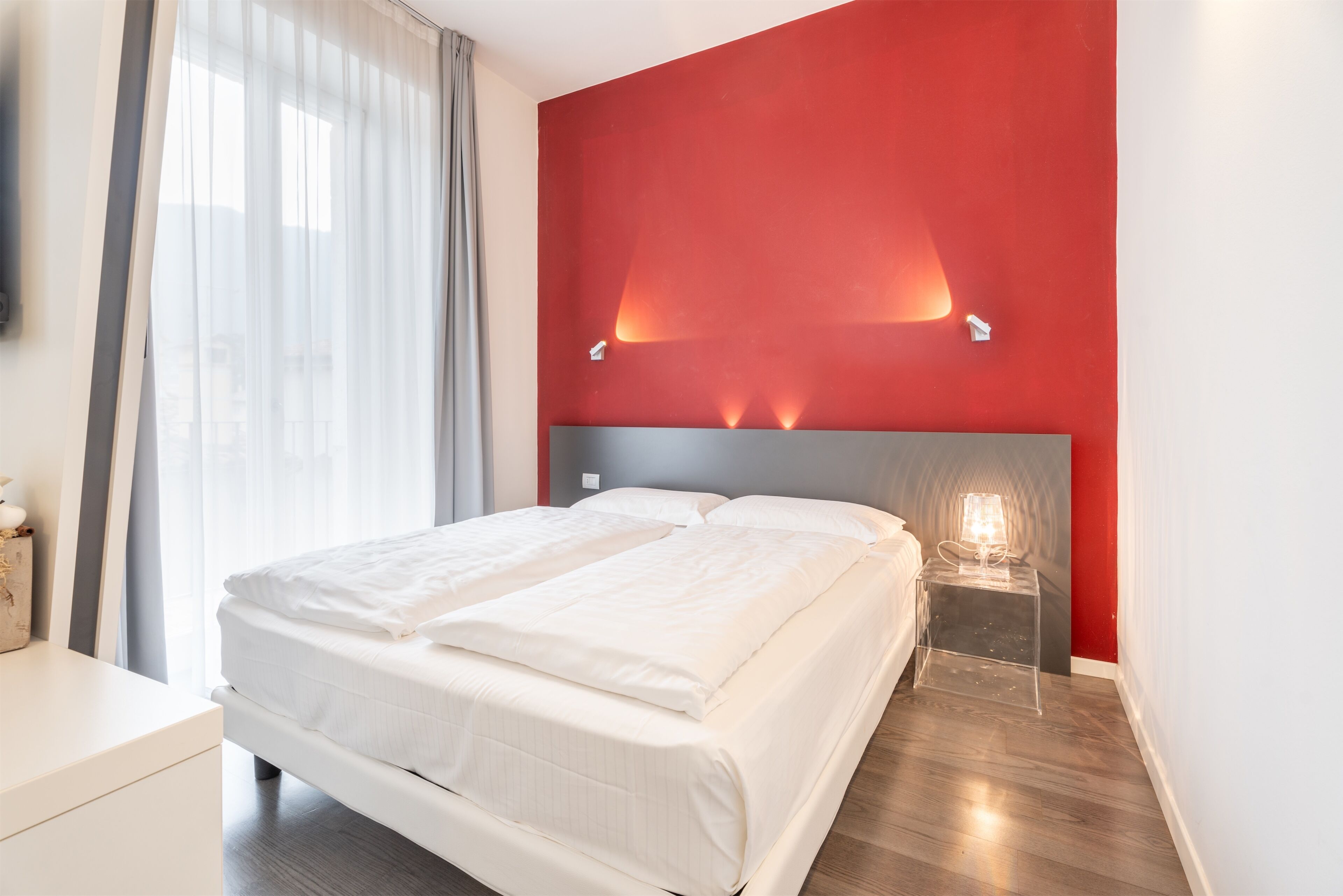 Habitación de hotel con cama, mesita de noche con lámpara y una pared de acento roja.