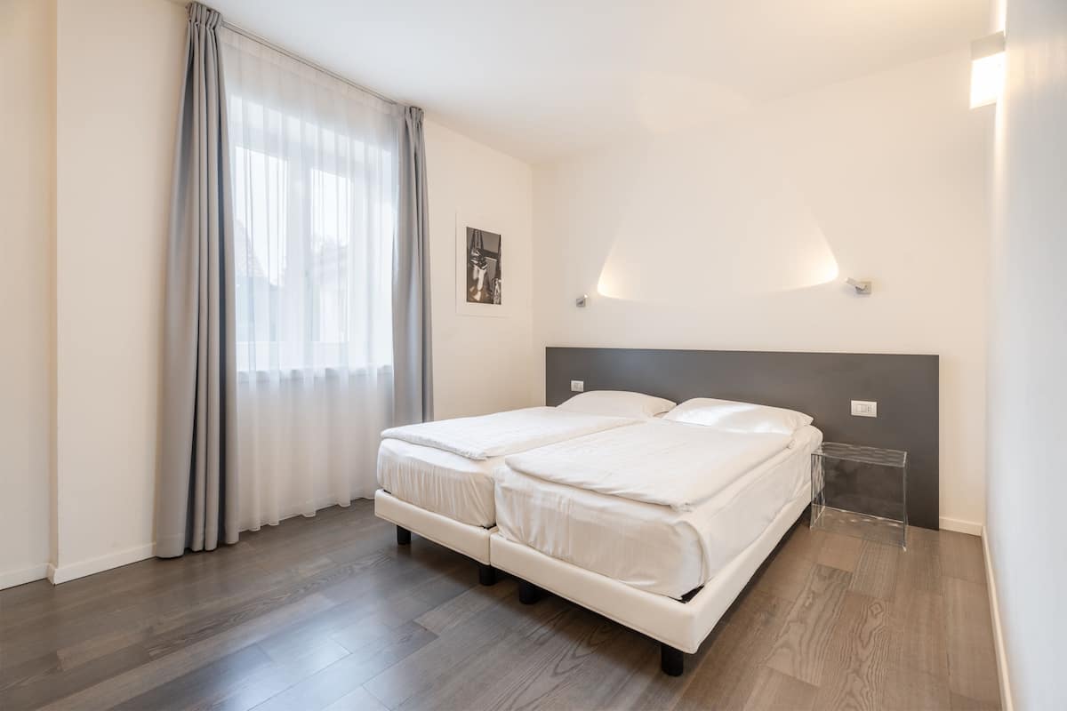 Ein modernes Schlafzimmer mit zwei Betten, einem großen Fenster mit transparenten Vorhängen und einem minimalistischen Design.