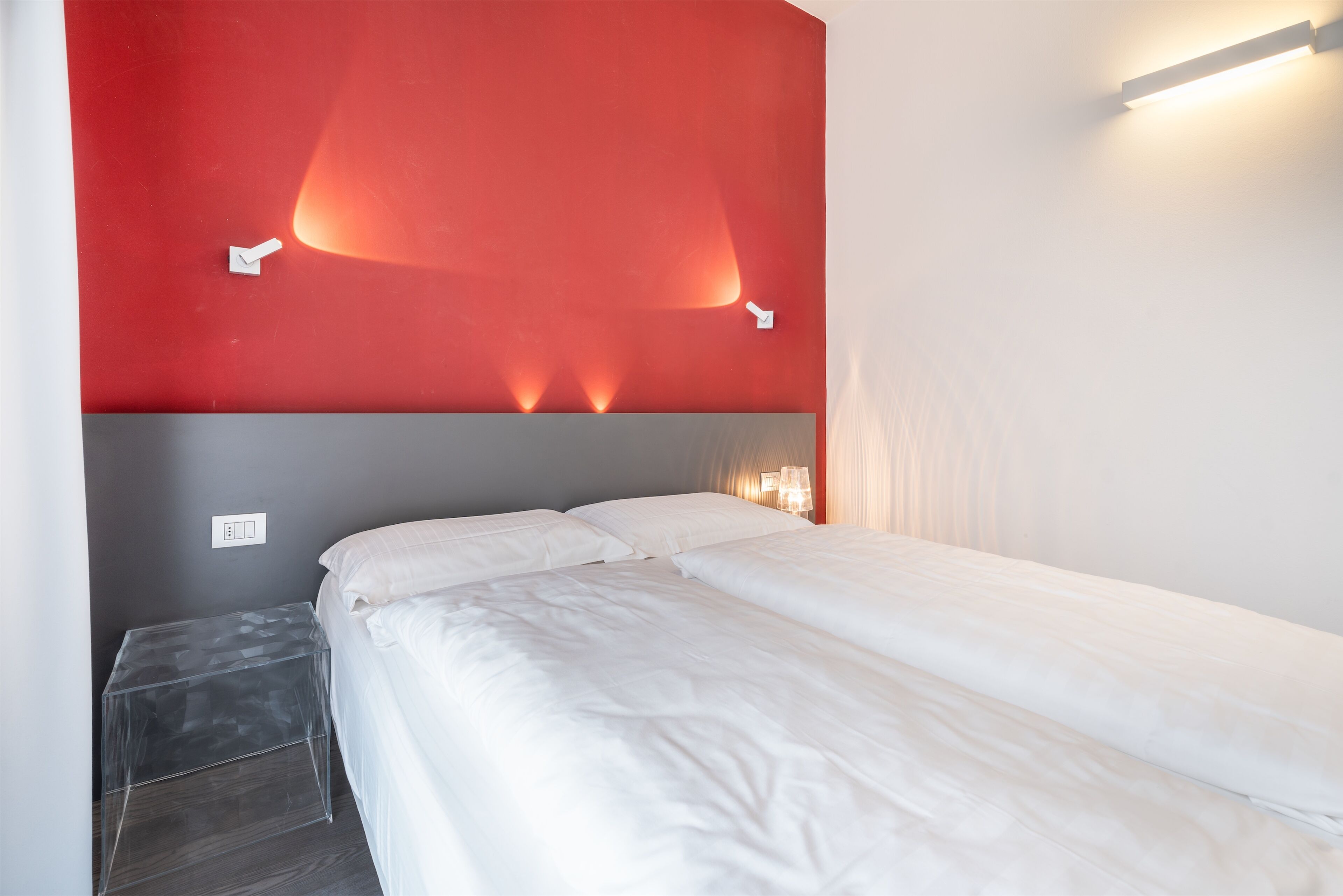 Habitación de hotel con cama, mesita de noche pequeña y una pared de acento roja con iluminación.