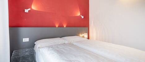 Habitación de hotel con cama, mesita de noche pequeña y una pared de acento roja con iluminación.