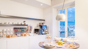 A breakfast buffet with various pastries and beverages, a coffee machine, and a view of the city through large windows. - Garnì On The Rock (Arco)