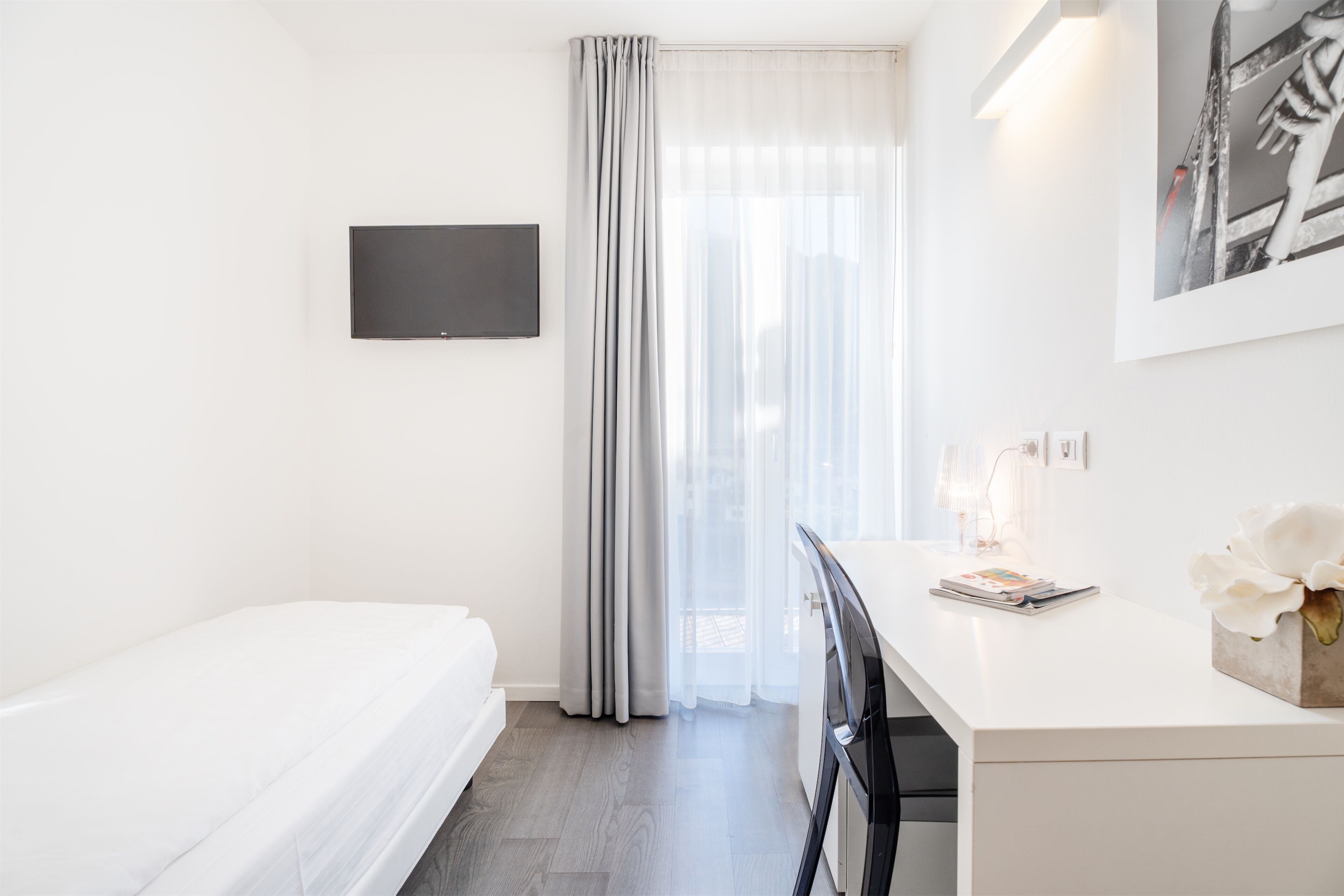 A modern hotel room with a bed, a desk with a chair, a flat-screen TV, a window with sheer curtains, and a framed picture on the wall.