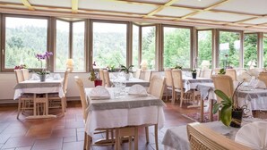 A restaurant with multiple tables, wicker chairs, and large windows offering a view of greenery. - Hotel Neuhauser (La Broque)