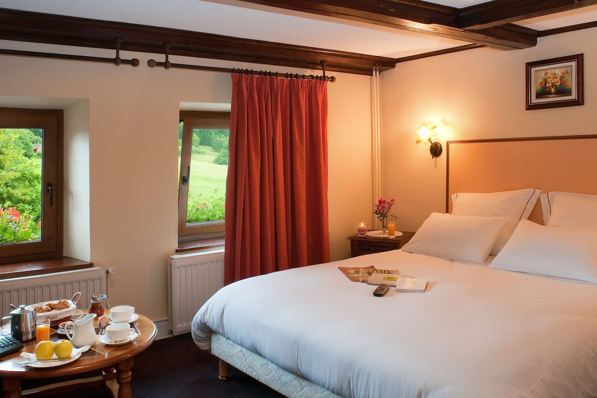Et hotelværelse med en seng, et bord med morgenmadsprodukter, et vindue med udsigt til grøn vegetation og en vægmonteret lampe.