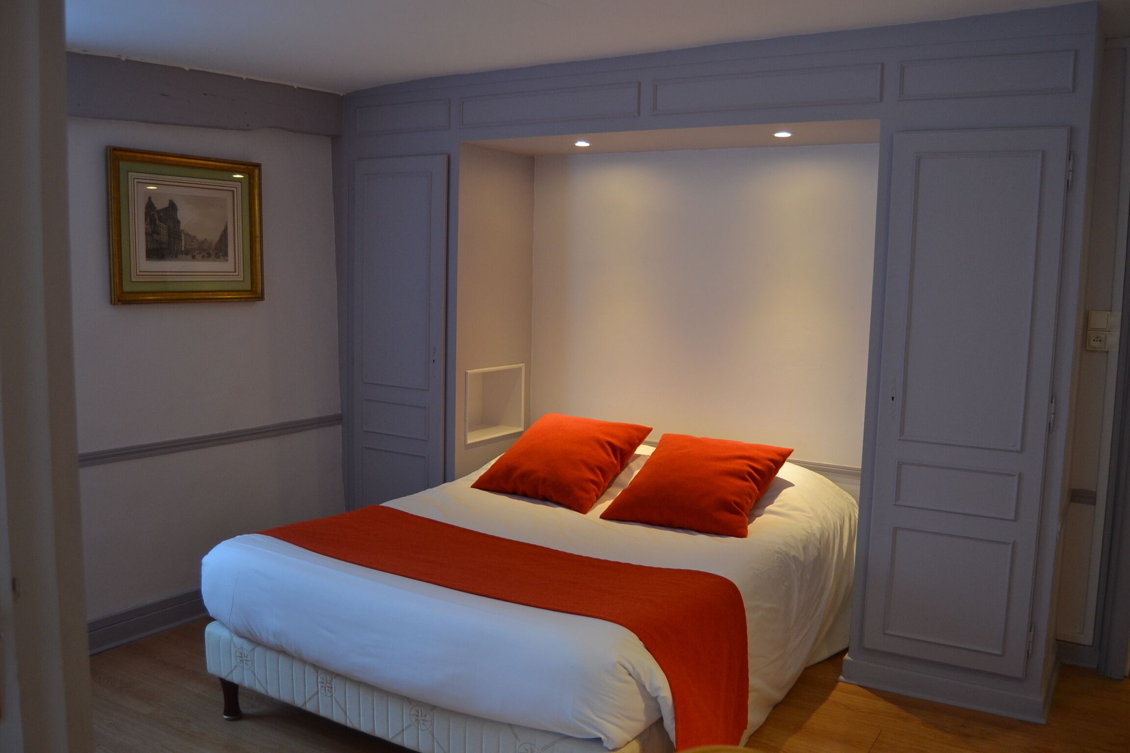 A neatly made bed with white linens and two orange pillows, a framed picture on the wall, and a door to the right.