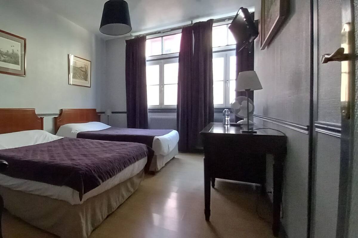 Ein Hotelzimmer mit zwei Einzelbetten, einem Schreibtisch mit Lampe, einem Stuhl und einem Fenster mit Vorhängen.