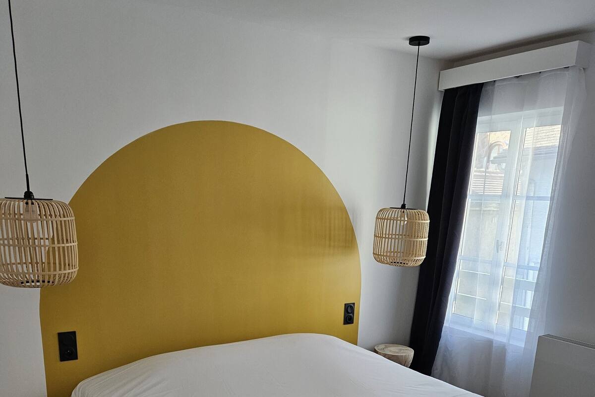 Ein Schlafzimmer mit einem großen Bett, einem gewölbten, gelben Kopfteil, zwei Nachttischen und einem Fenster mit transparenten Vorhängen.