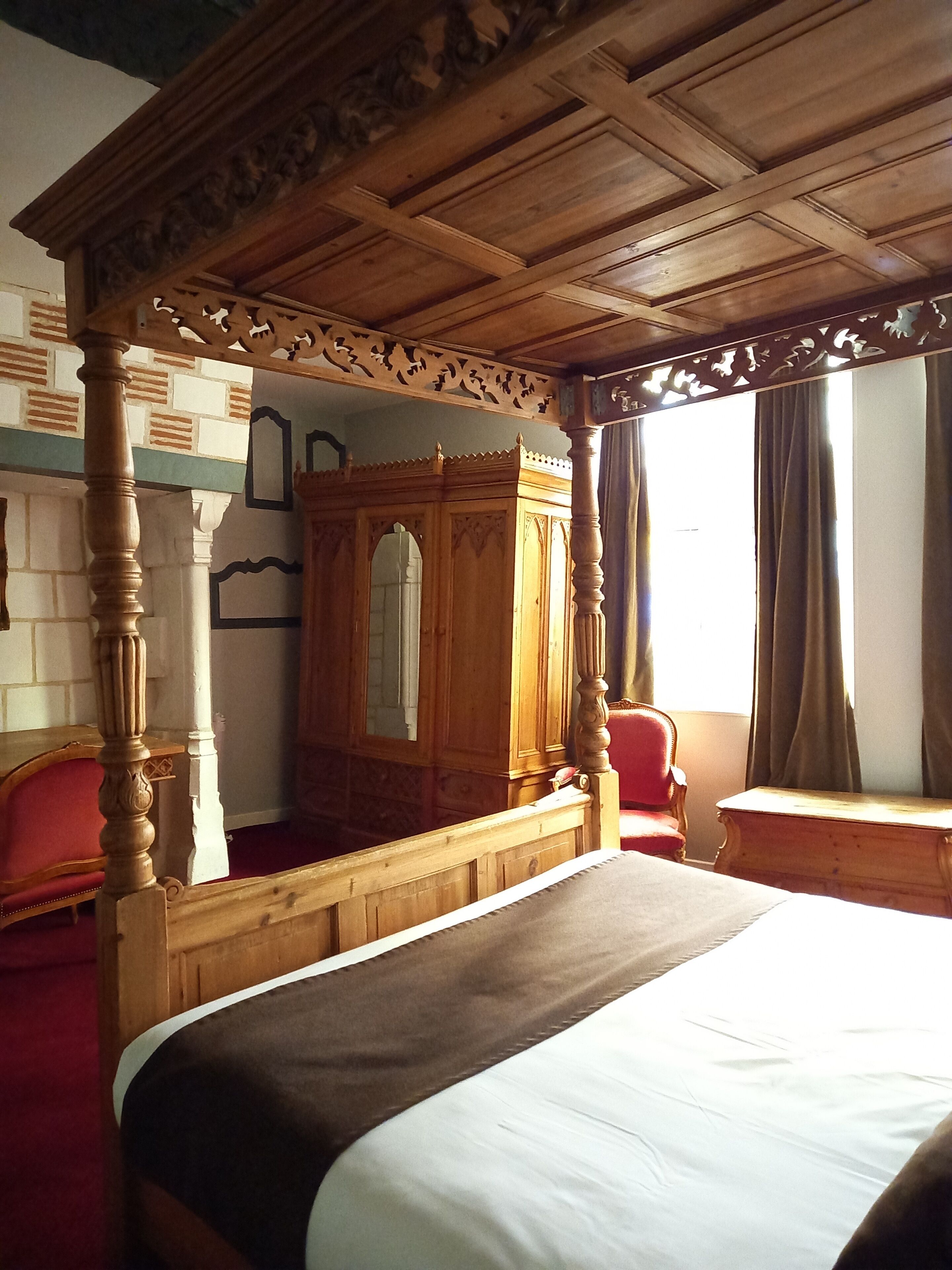 Une chambre à coucher avec un lit à baldaquin, une armoire en bois, un fauteuil rouge et une fenêtre avec des rideaux.