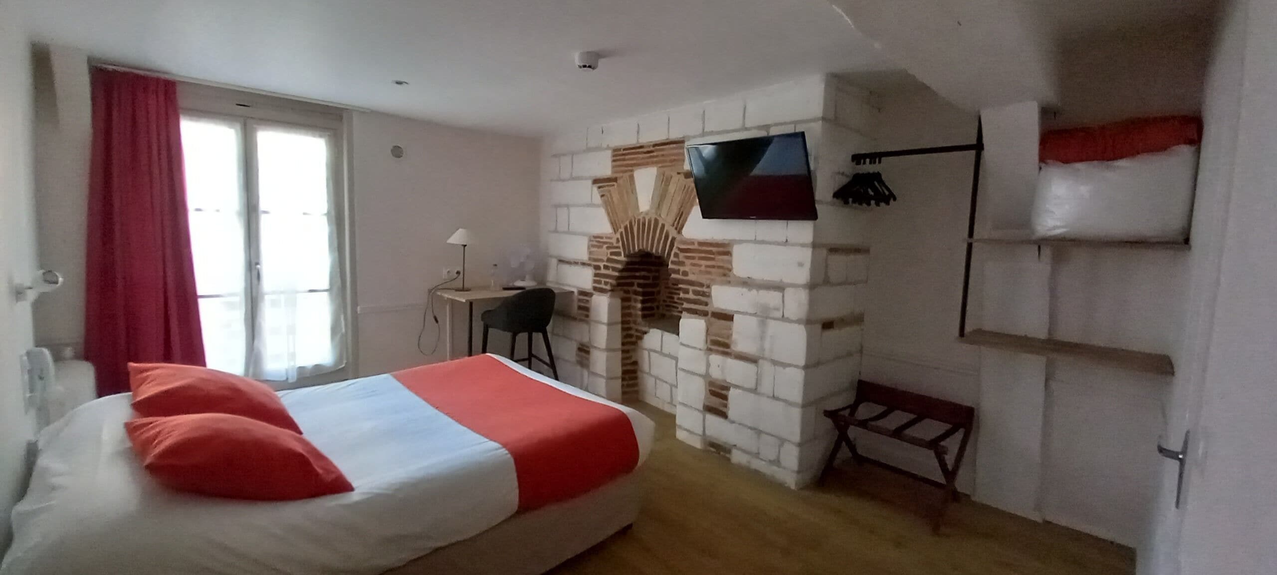 A bedroom with a bed, a TV mounted on a brick wall, a small table, and a chair.
