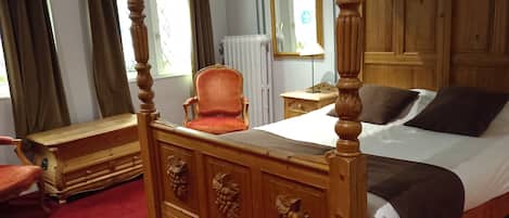 A four-poster bed with carved wooden details, a red upholstered chair, and a wooden nightstand with a lamp.