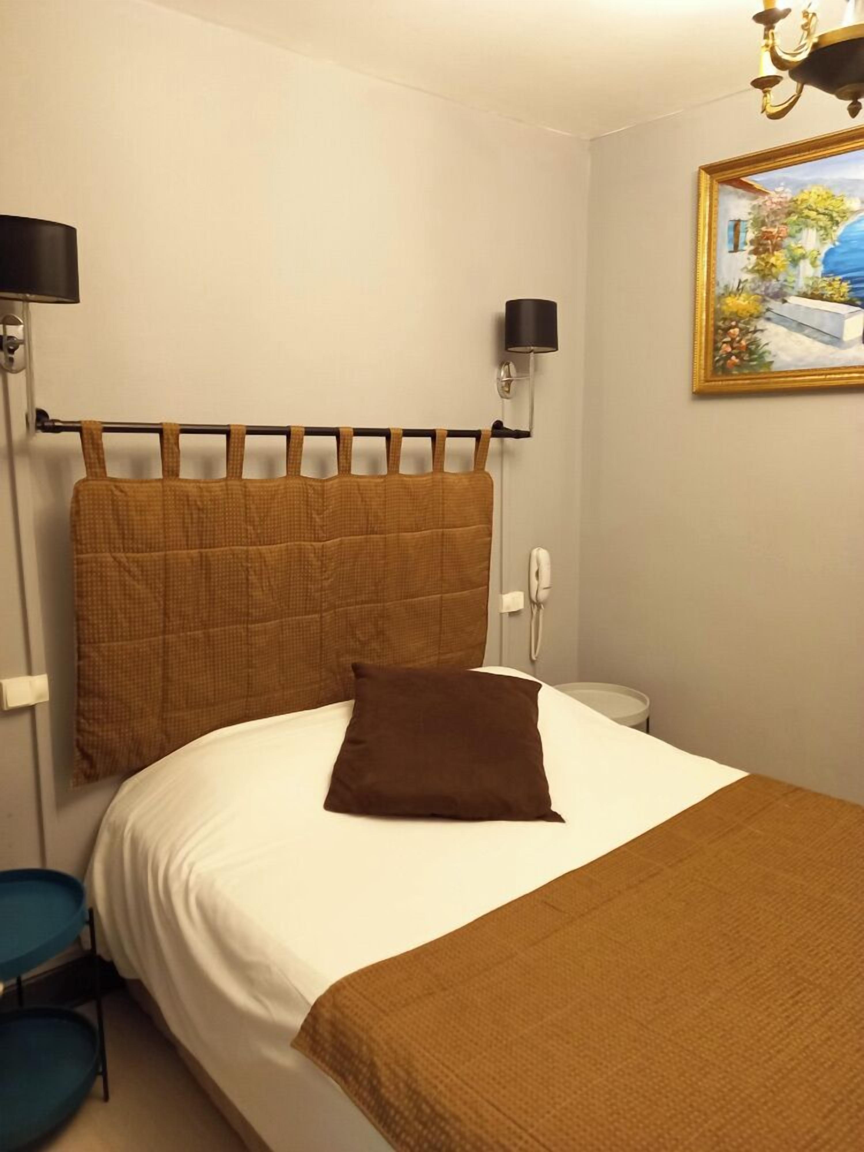Un letto ben rifatto con una testiera marrone, un cuscino marrone e una coperta bianca. C'è un comodino con una lampada, un telefono fissato al muro e un quadro incorniciato sopra il letto.