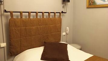 A neatly made bed with a brown headboard, a brown pillow, and a white bedspread. There is a small bedside table with a lamp, a wall-mounted phone, and a framed painting above the bed.