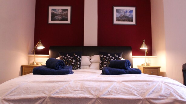 A neatly made bed with white linens and two blue towels, a wooden nightstand with a lamp, and two framed pictures on the wall. - Acomb Lodge (Harrogate)