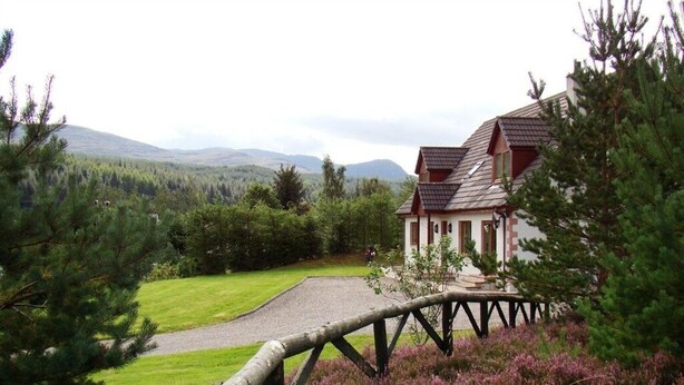 A house with a stone foundation, a red roof, and a wooden fence in a natural setting with heather and pine trees. - Binnilidh Mhor B&B (Inverness)