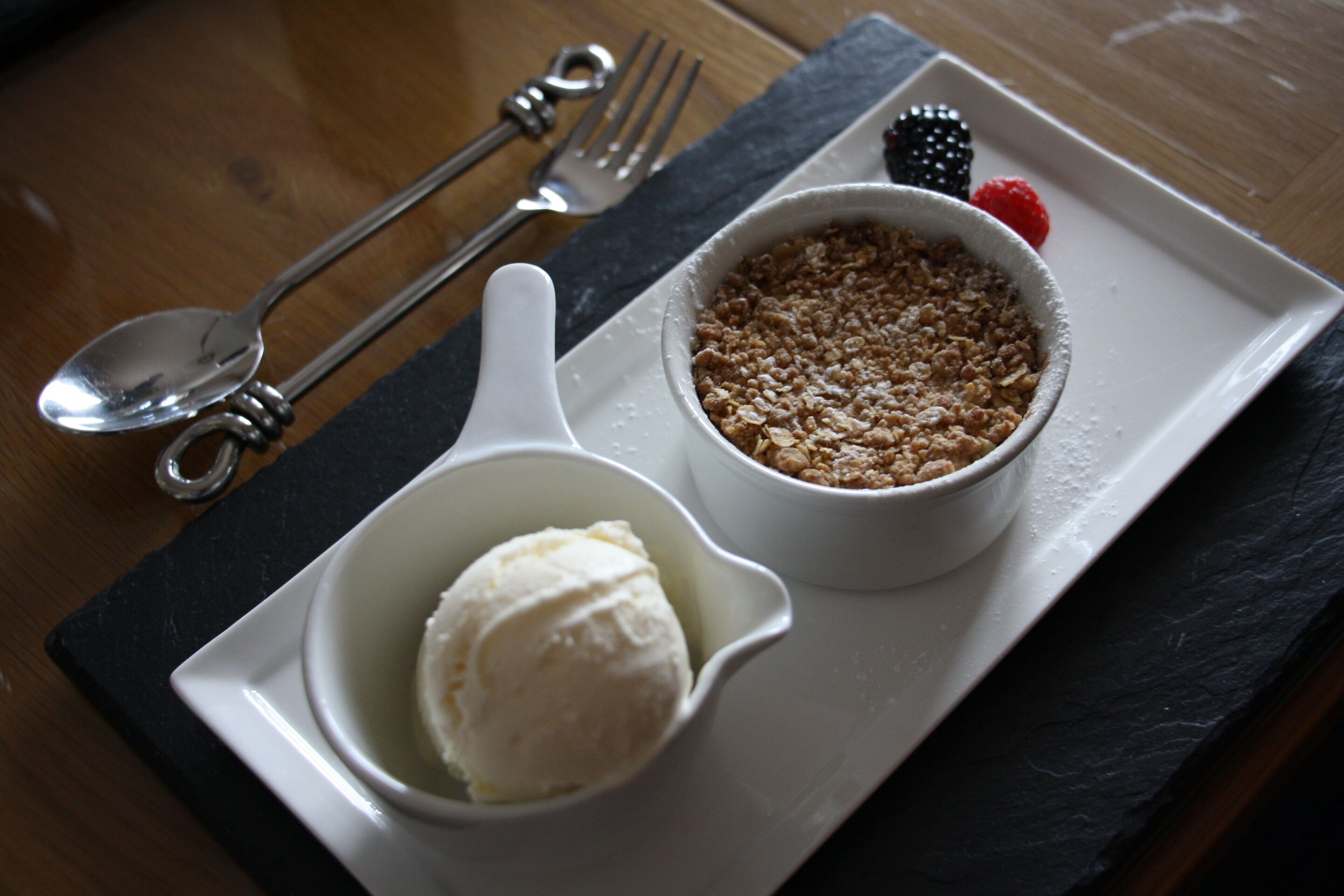 A dessert plate with ice cream and a crumble dessert, accompanied by a fork and spoon on a slate serving board.