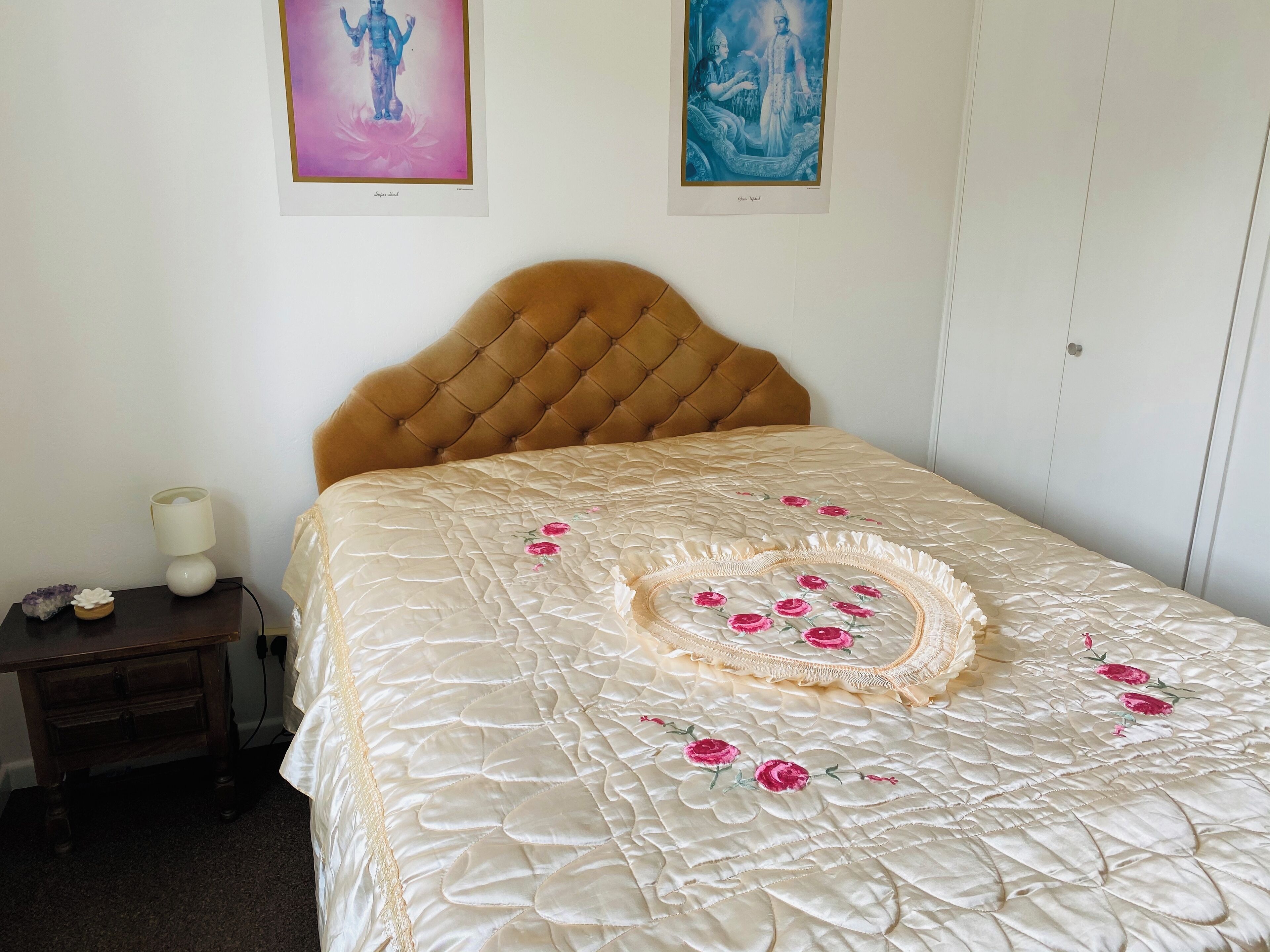 A bedroom with a tufted headboard, a quilted bedspread with floral embroidery, a bedside table with a lamp, and two framed pictures on the wall.