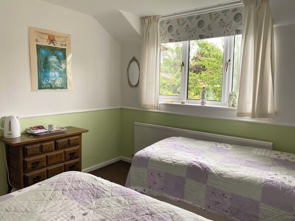 A bedroom with two single beds, a wooden dresser, a radiator, a window with curtains, and a wall decoration.