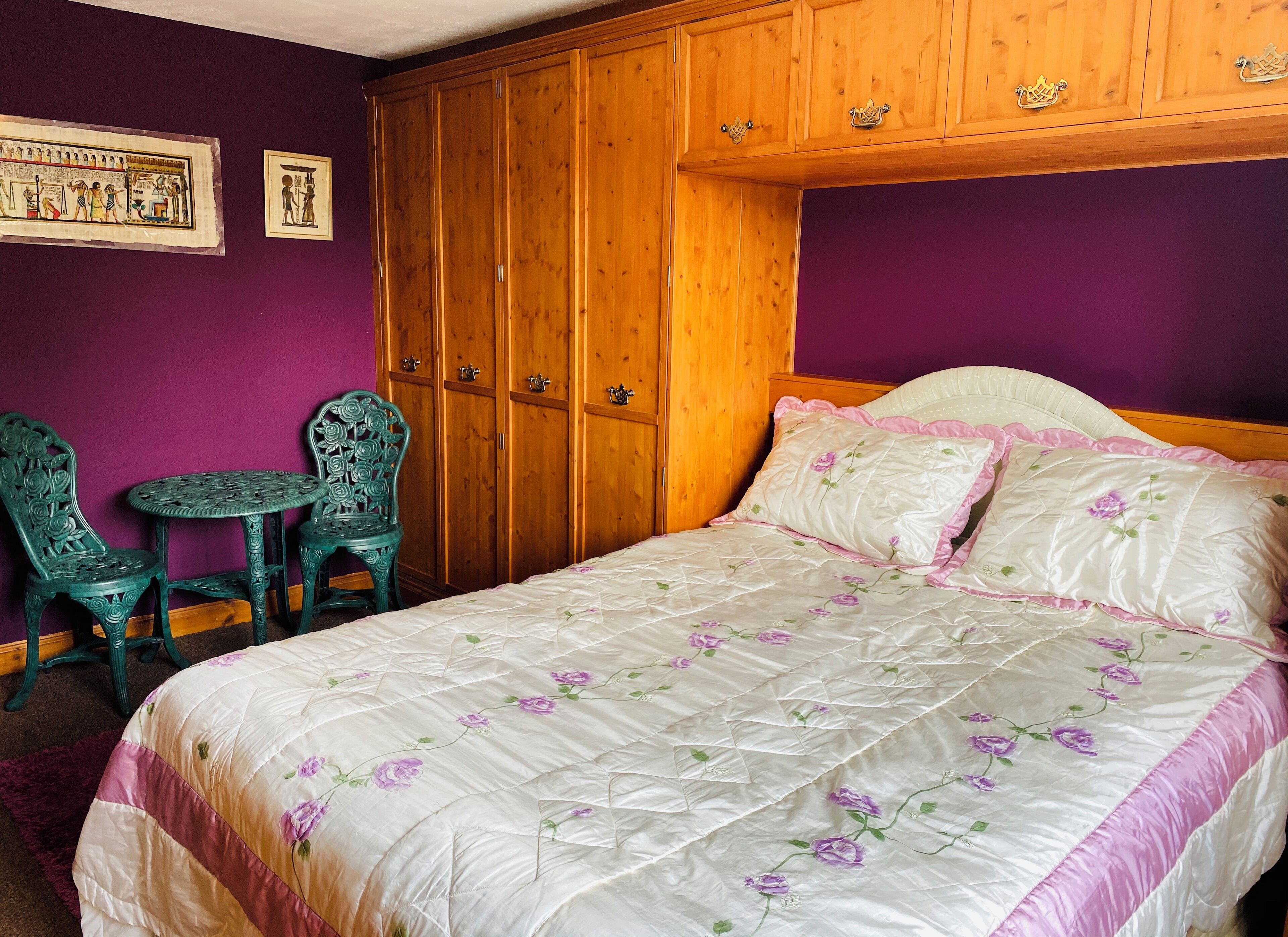 Un dormitorio con cama, armario de madera, dos sillas verdes y una mesita.