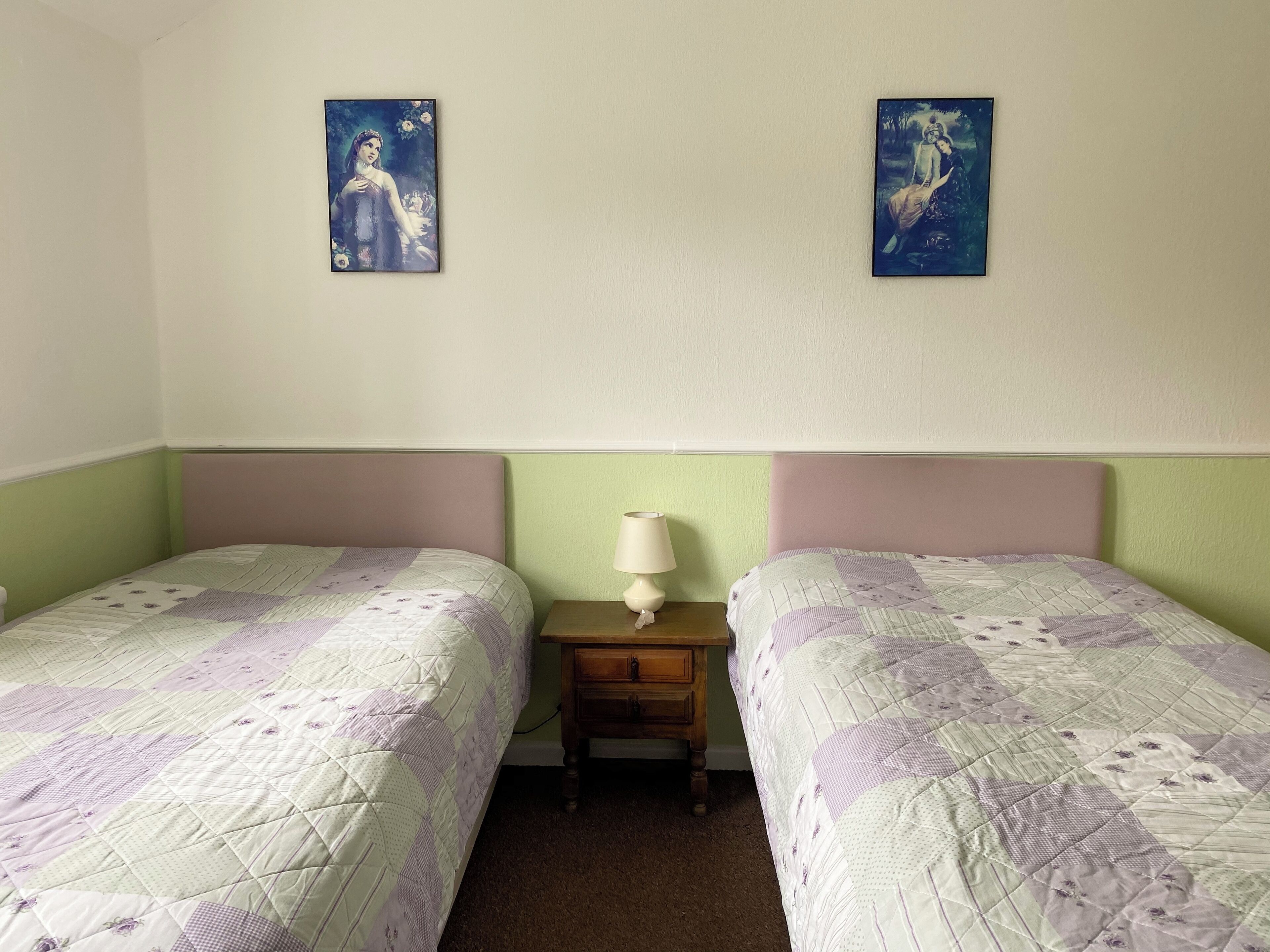 A room with two beds, each with a floral quilt, a small bedside table with a lamp, and two paintings on the wall.