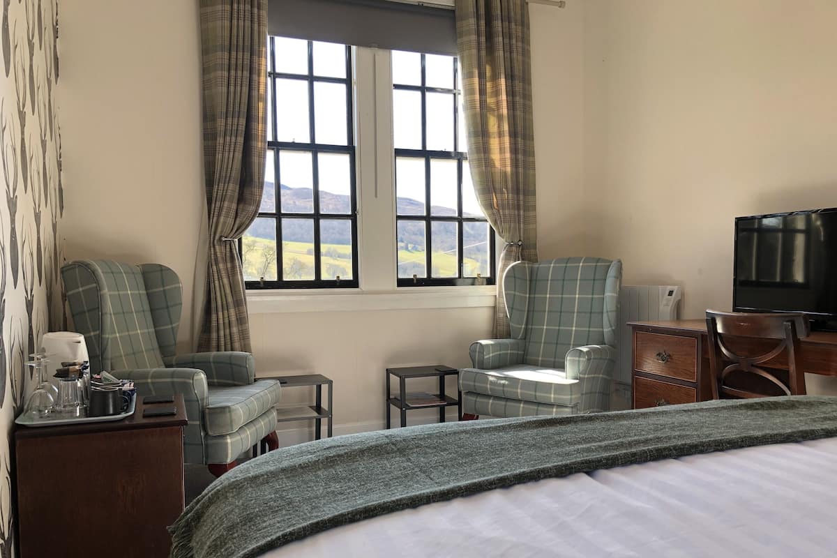 Une chambre d’hôtel comprenant un lit, deux fauteuils, un bureau avec une télévision et une fenêtre avec des rideaux.