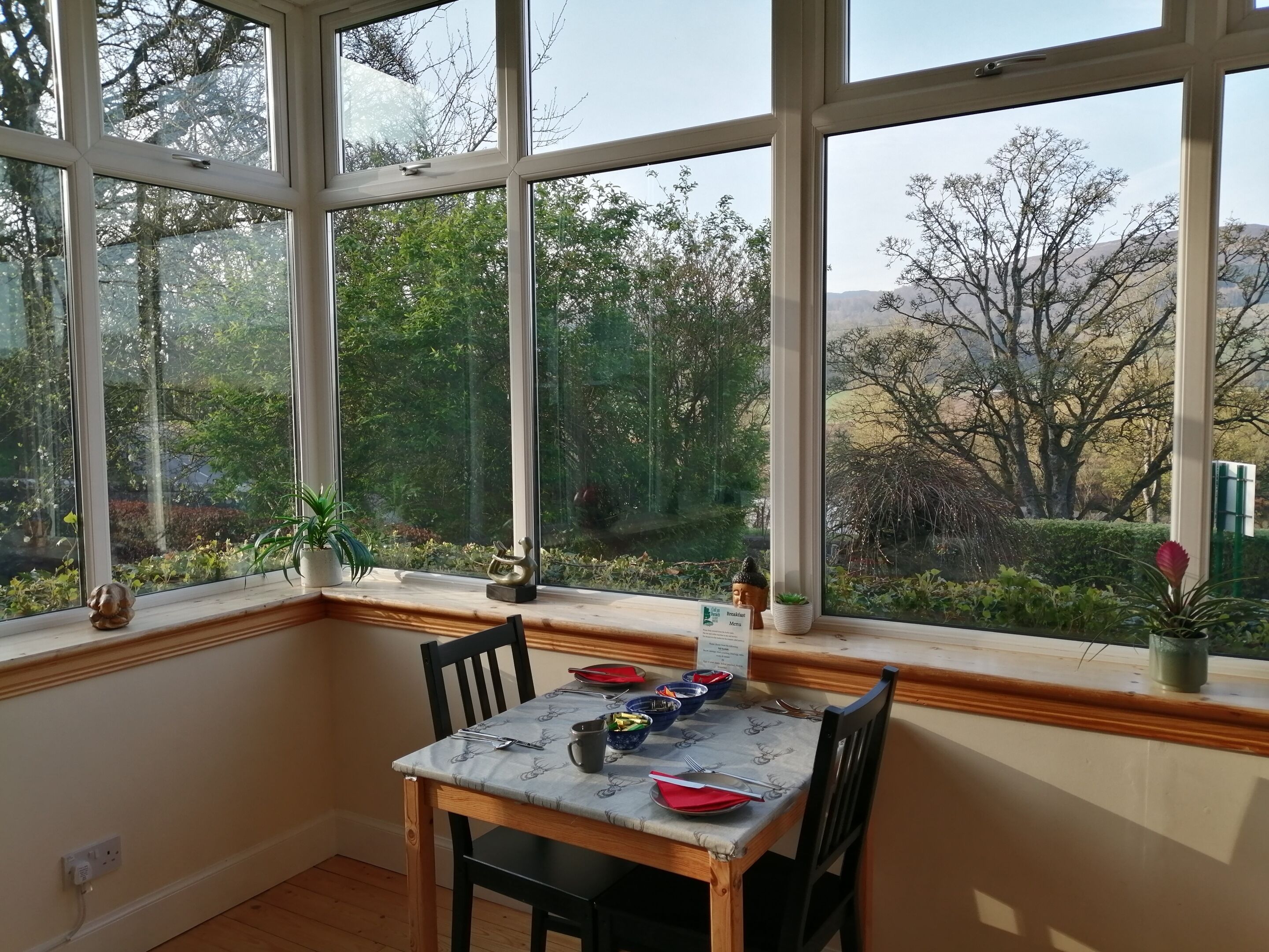 Un coin repas avec une table dressée pour deux personnes, une fenêtre donnant sur des arbres et des collines, et une petite statue de Bouddha posée sur le rebord de la fenêtre.