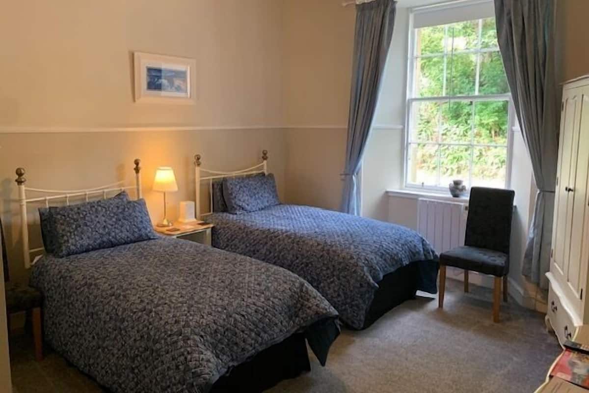 Une chambre avec deux lits simples, une table de chevet avec une lampe, une chaise, une fenêtre avec des rideaux et un radiateur.