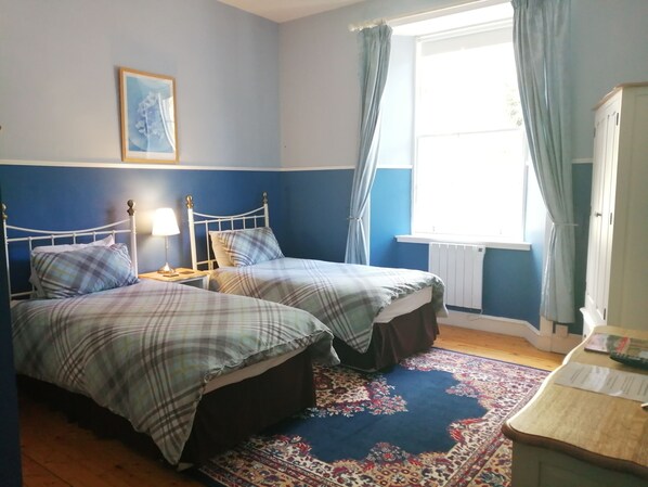 A bedroom with two beds, a nightstand, a window with curtains, a radiator, and a small table with a lamp. - Cuil an Daraich Guest House (Pitlochry)