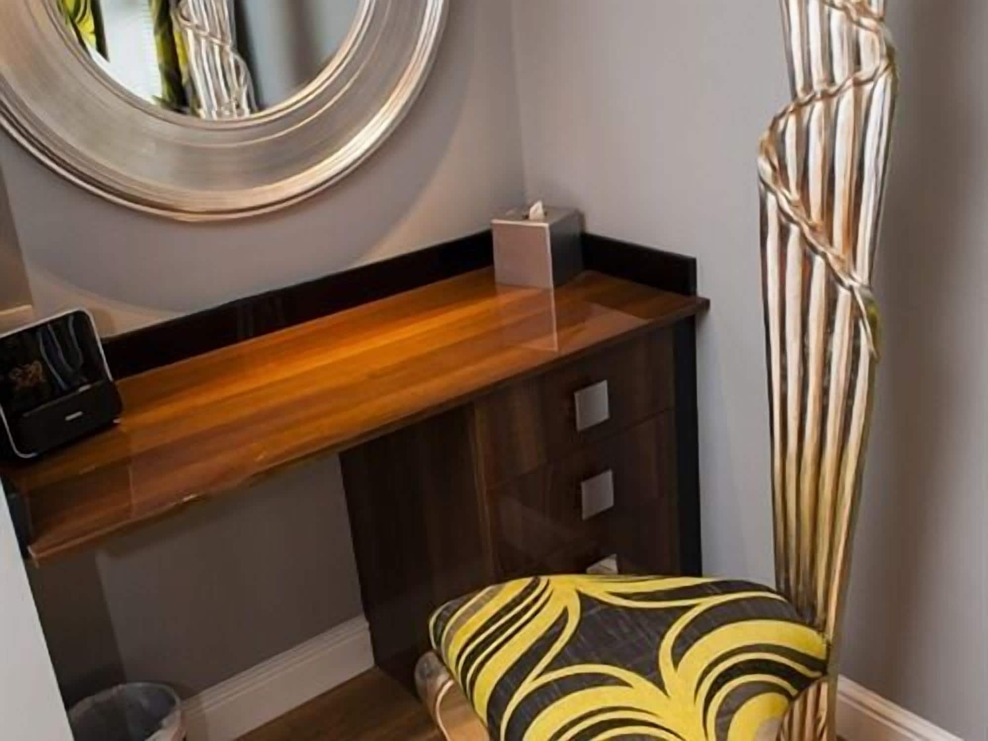 A compact room with a wooden desk, a chair with a striped cushion, a small trash bin, and a mirror on the wall.