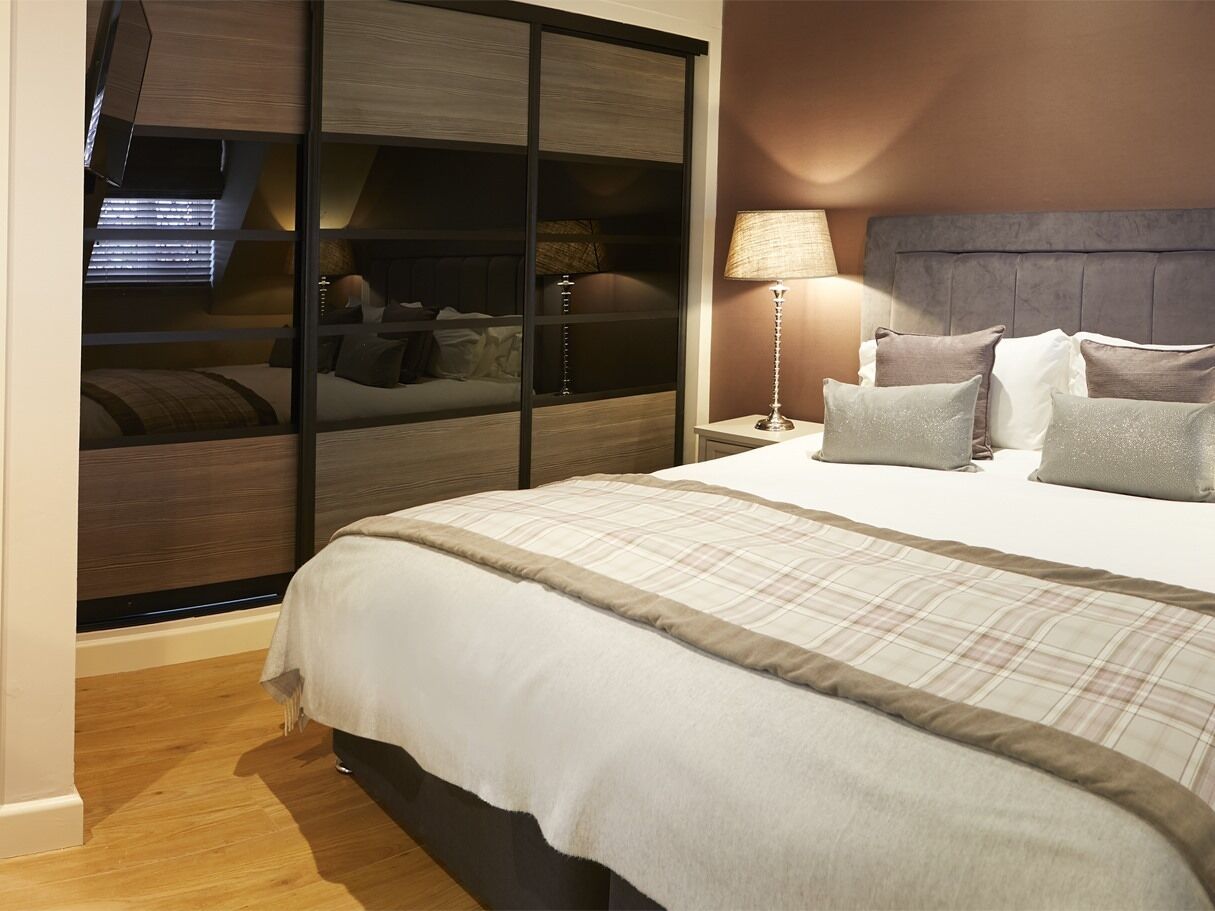Ein modernes Schlafzimmer mit einem großen Bett, einem Schiebekleiderschrank, einem Nachttisch mit Lampe und Blick auf eine Stadtlandschaft durch ein Fenster.