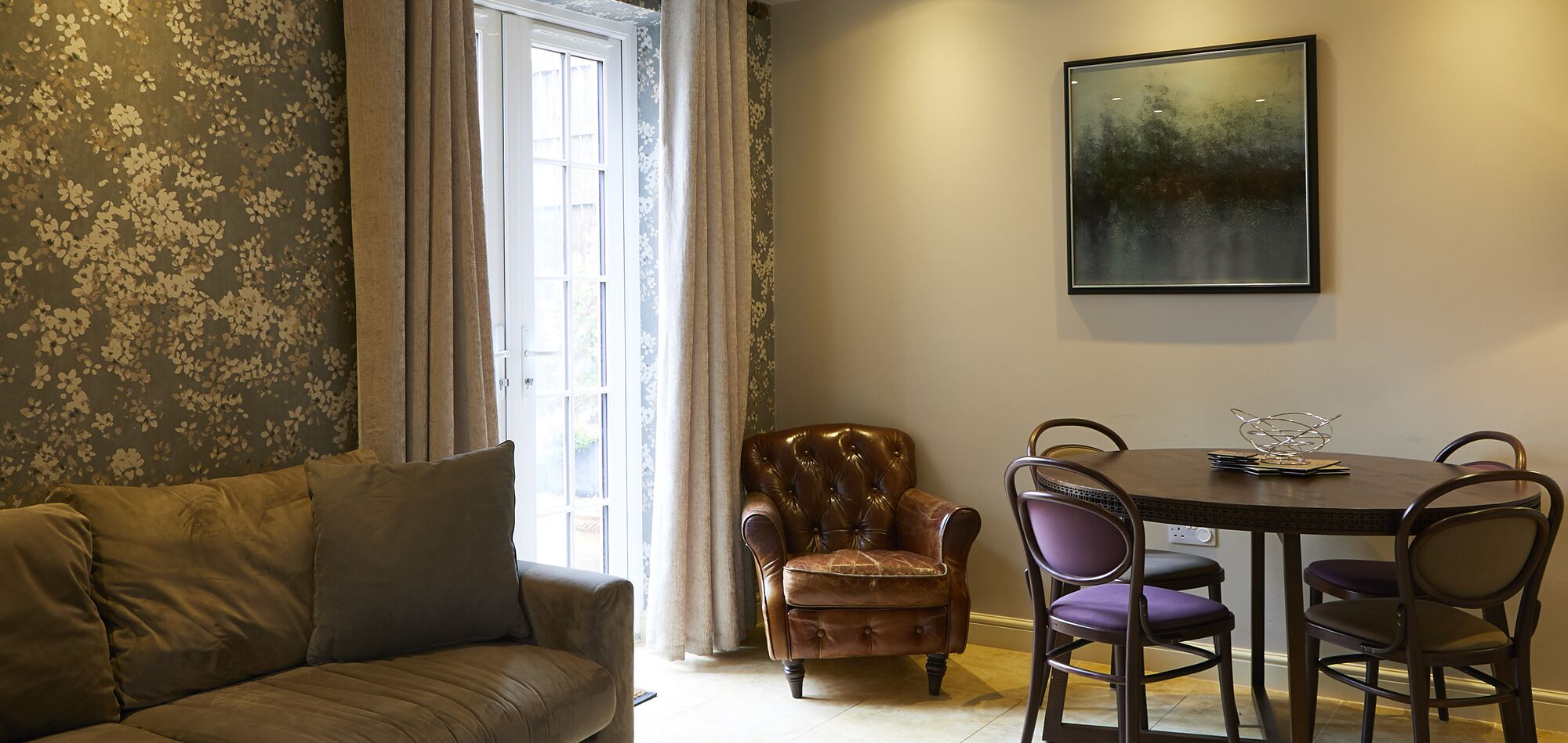A living room with a brown leather sofa, a tufted leather armchair, and a round dining table with four chairs. There is a large painting on the wall and a window with curtains.