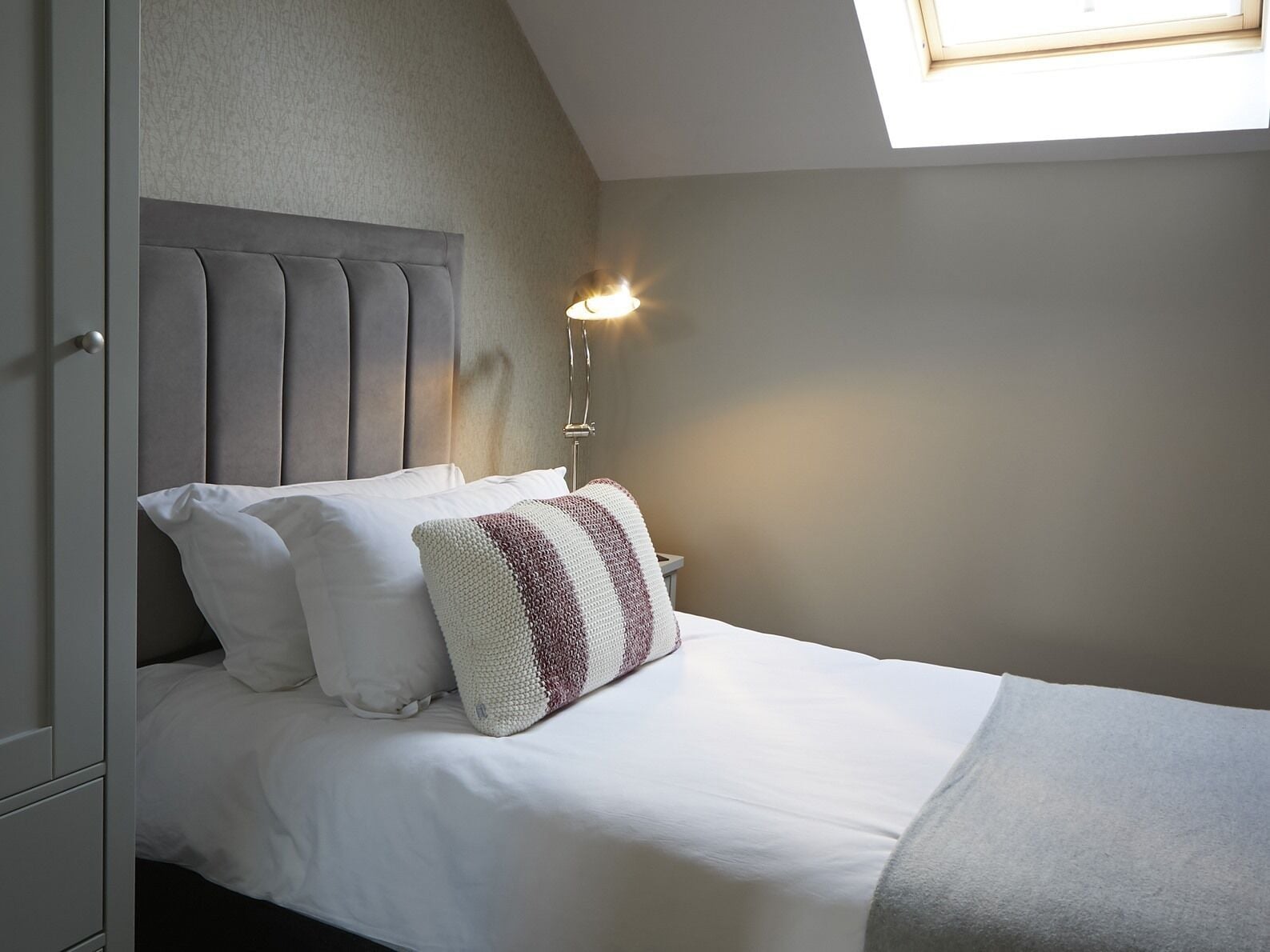 A neatly made bed with white linens and a grey headboard, a bedside lamp, and a window with natural light.