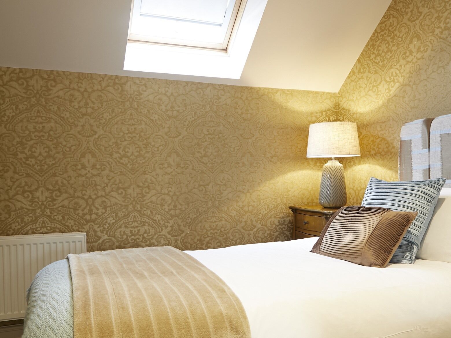 A bedroom with a skylight, patterned wallpaper, a bed with a striped blanket, a bedside lamp, and a nightstand with a lamp.