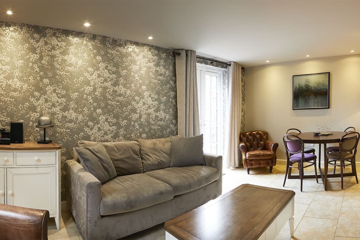 Een woonkamer met een grijze bank, een bruine leren fauteuil, een houten salontafel, een eettafel met stoelen, en een wit kastje met een ingelijst diploma.