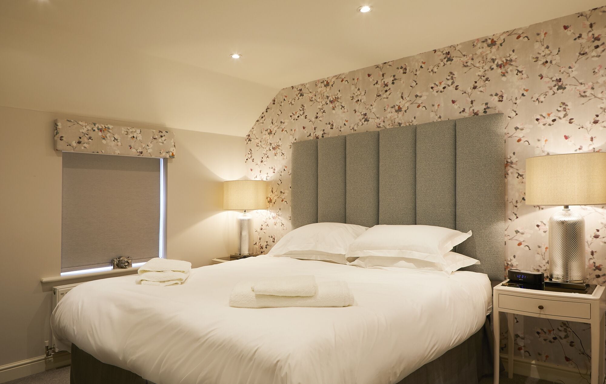 Una cama bien hecha con sábanas y almohadas blancas, una mesita de noche con una lámpara y un estampado floral en el papel tapiz de las paredes.