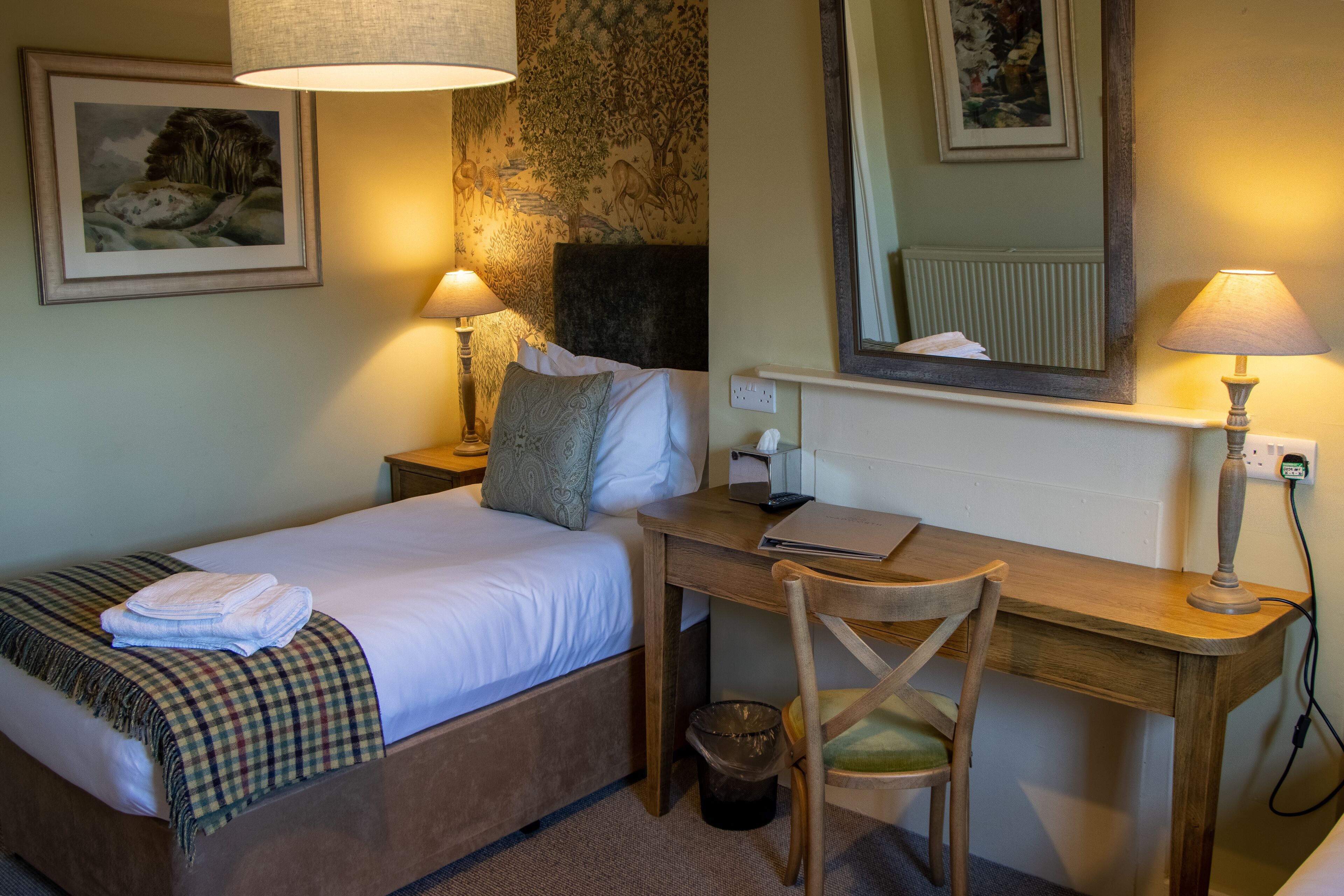 A hotel room with a bed, desk, chair, lamp, and a wall with a painting.