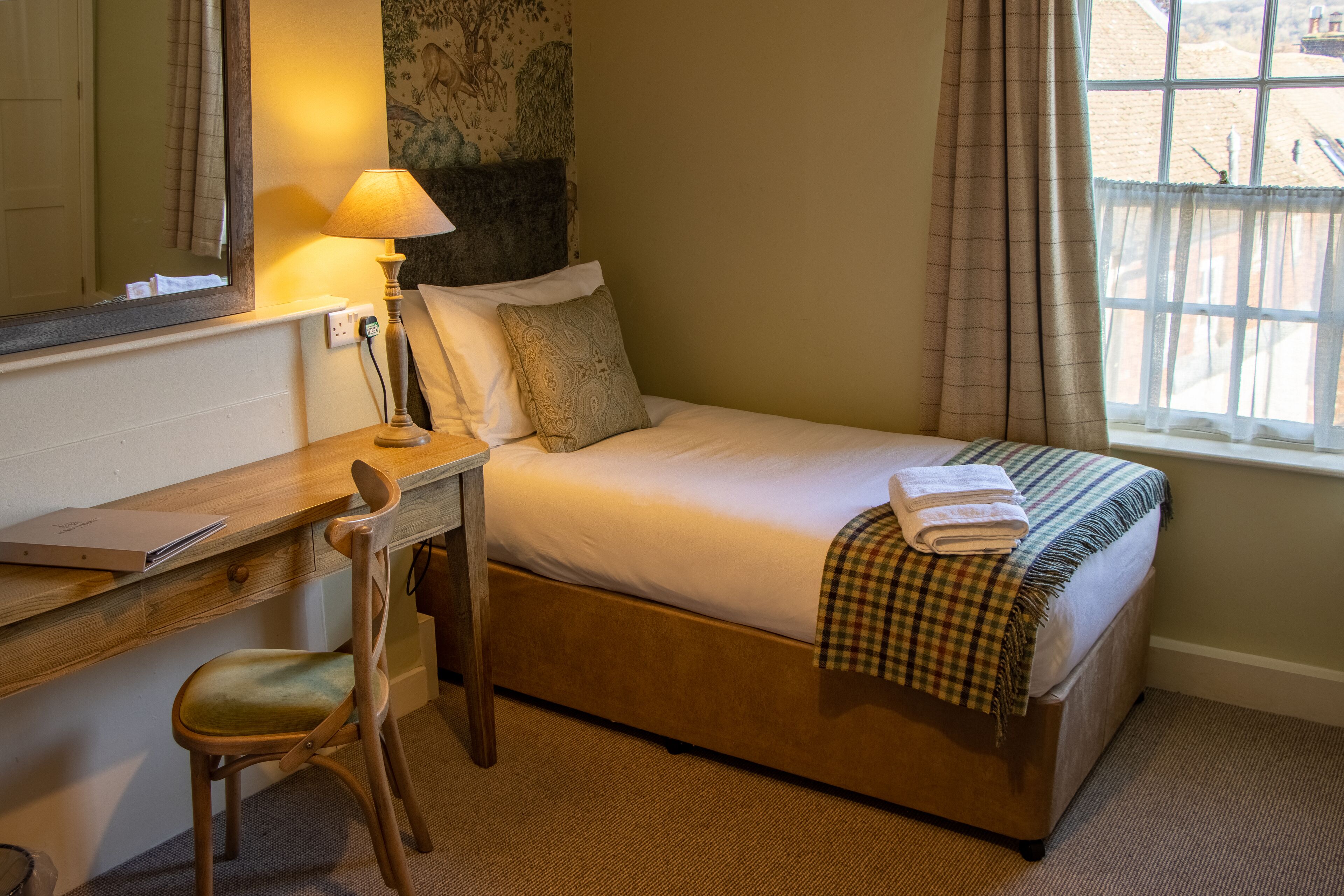 A hotel room with a single bed, a desk with a chair, a lamp, and a window with curtains.