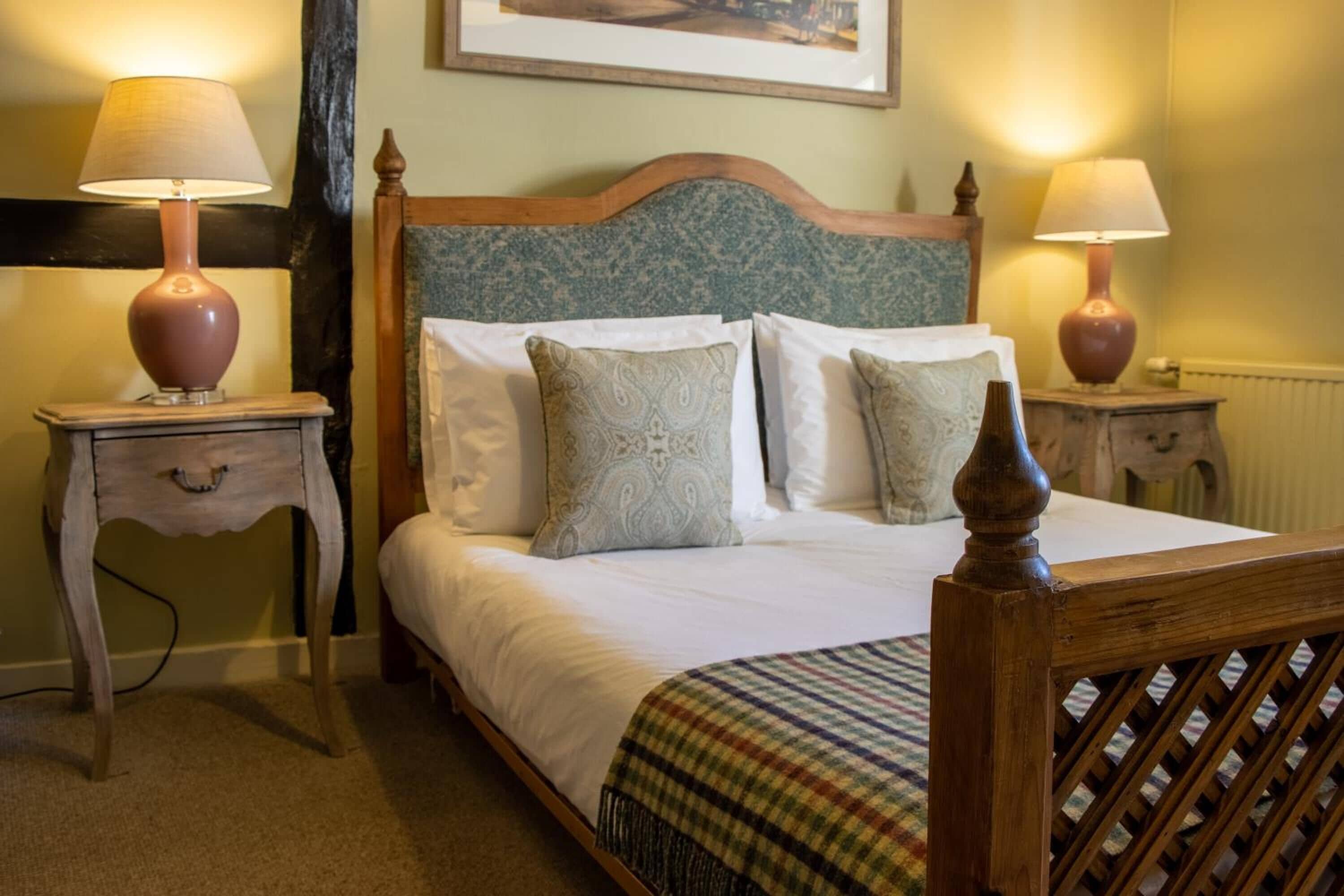 A neatly made bed with white linens and patterned pillows, flanked by two bedside tables with lamps, in a room with a wooden headboard and a framed picture above the bed.