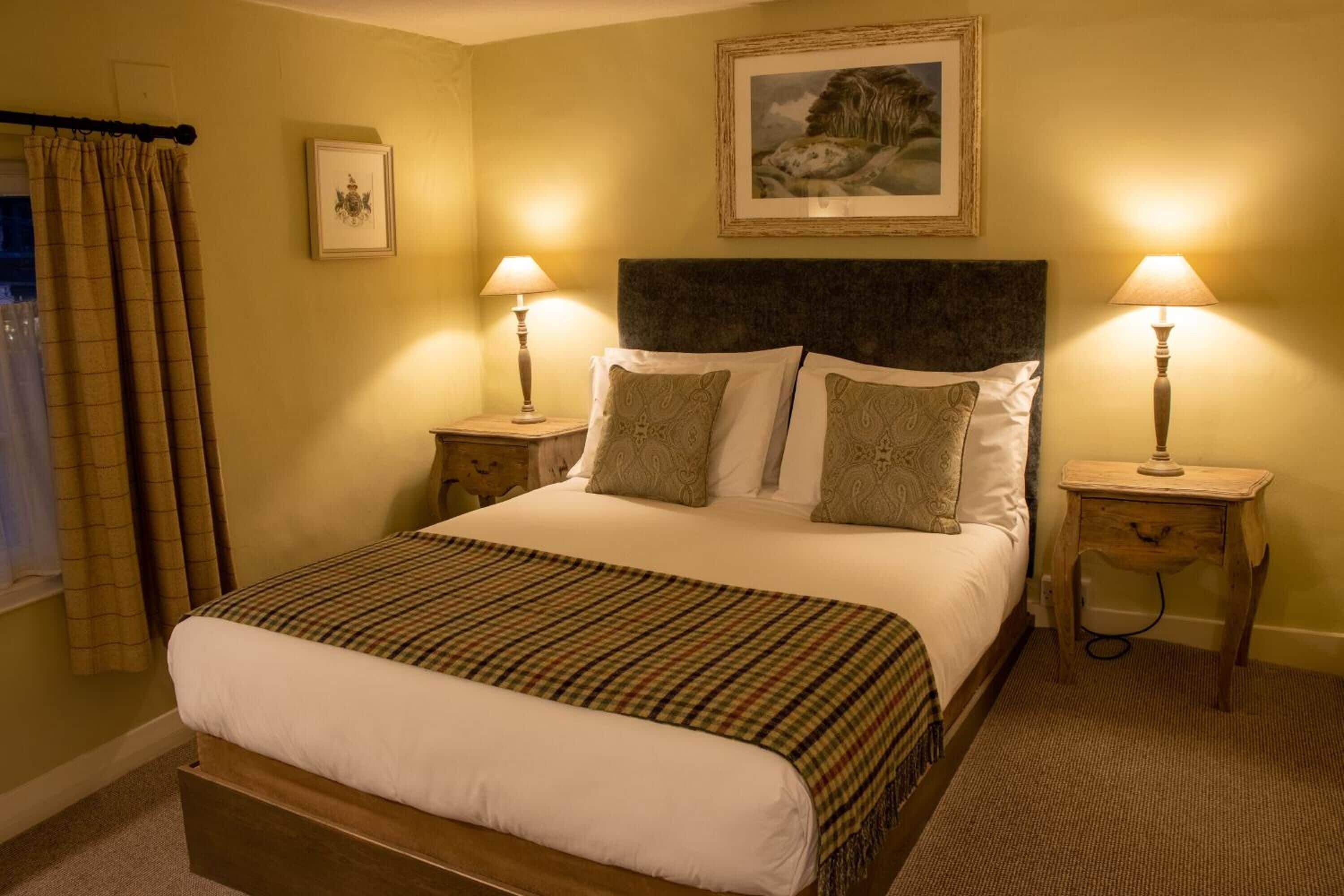 Una cama bien hecha con colcha escocesa, dos lámparas de noche, un cuadro enmarcado sobre la cama y una ventana con cortinas.