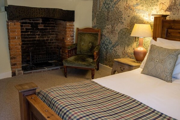 A bedroom with a brick fireplace, a green armchair, a bedside table with a lamp, and a bed with a plaid blanket.
