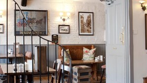 Un interior rústico con vigas de madera a la vista, una escalera, una zona de comedor con sillas de madera, un banco y una puerta blanca con lámpara.