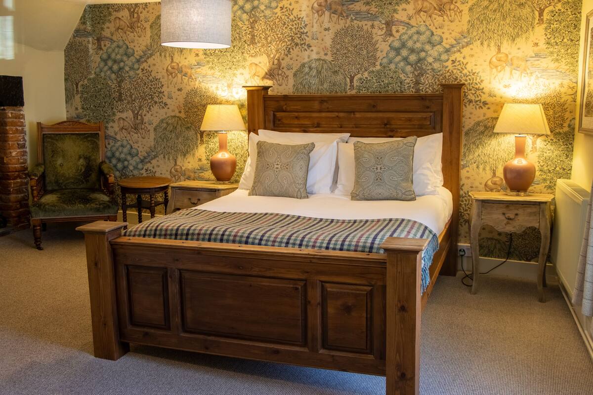 A wooden bed with a plaid bedspread, flanked by two nightstands with lamps, a chair, and a wall with a nature-themed mural.