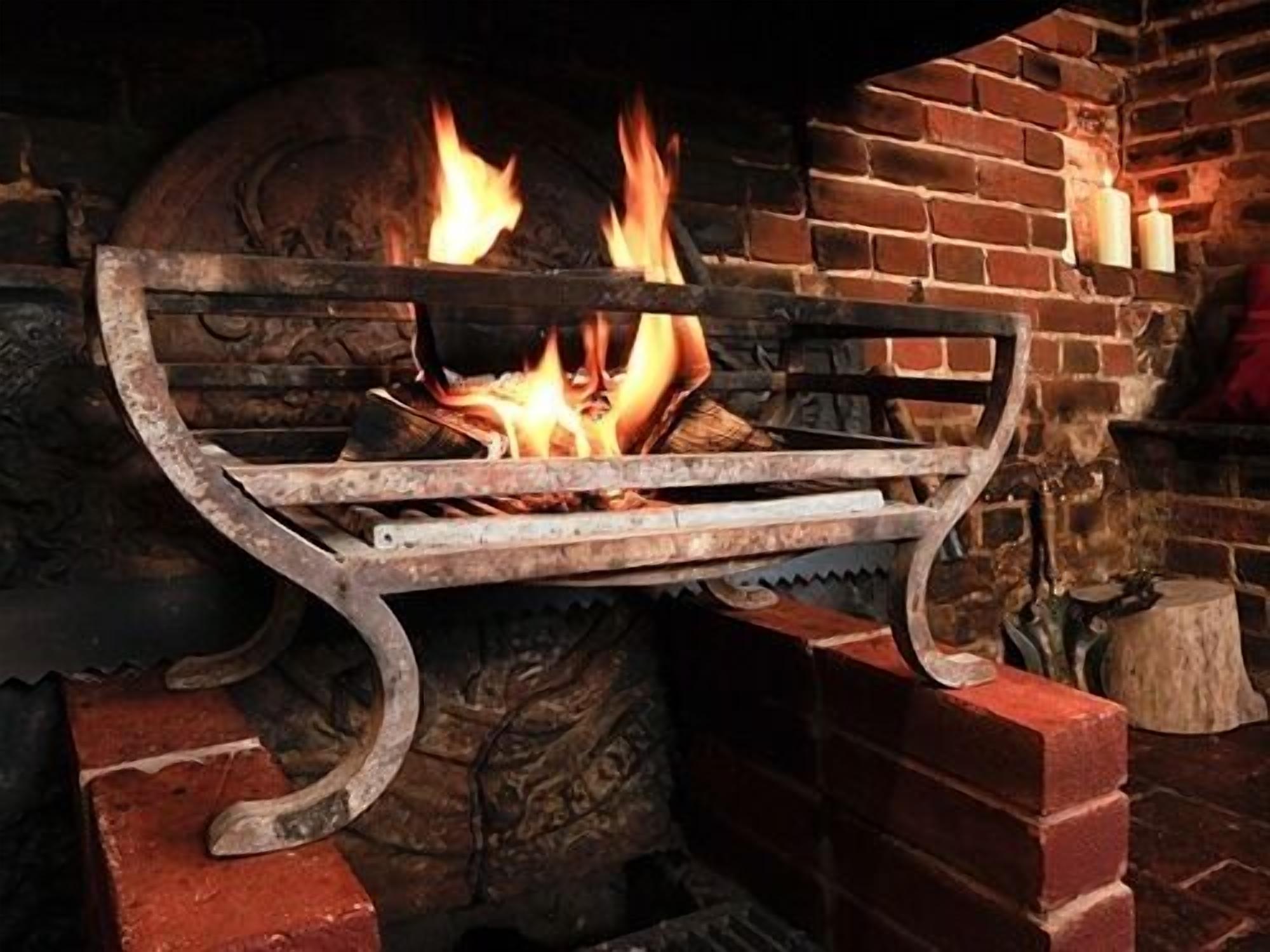 A fireplace with a metal grate, burning logs, and a brick wall.